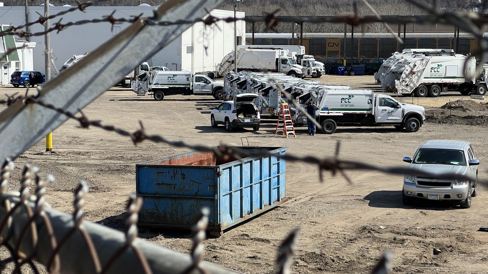 A barbed wire fence surrounding a dirt parking lot where white trash collection trucks are parked