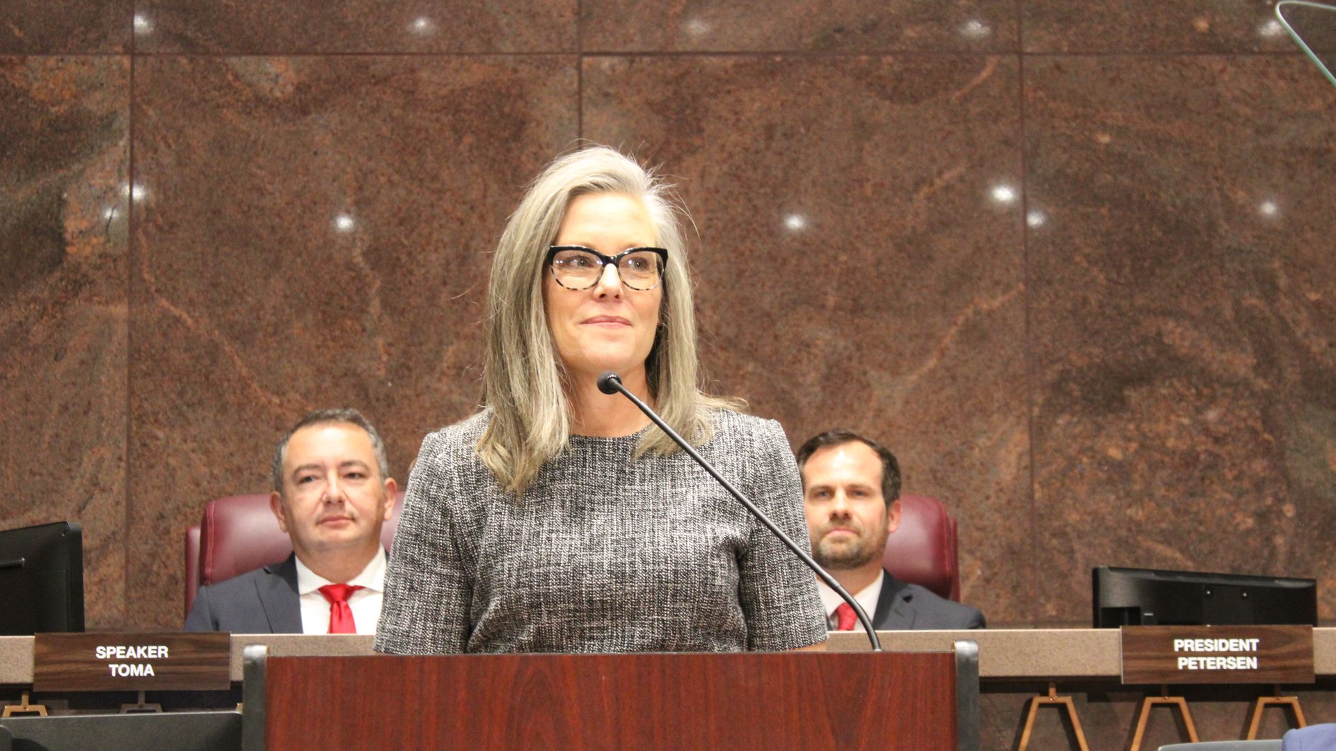 Arizona Governor Katie Hobbs stands at a lectern with House Speaker Ben Toma and Senate President Warren Petersen seated behind her.