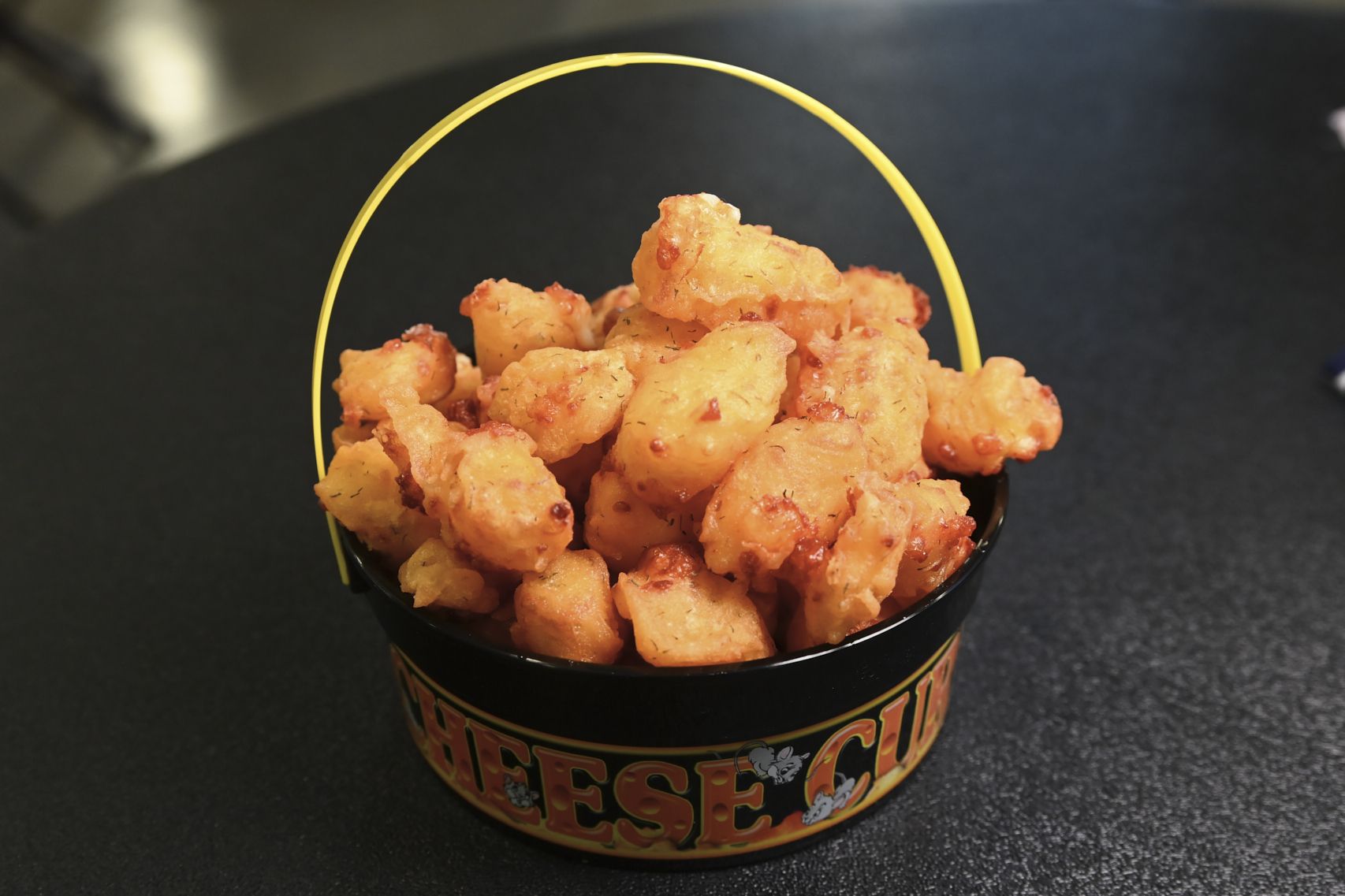 Crispy orange fried chicken bites piled in a black bucket with a yellow curved handle on a dark countertop.
