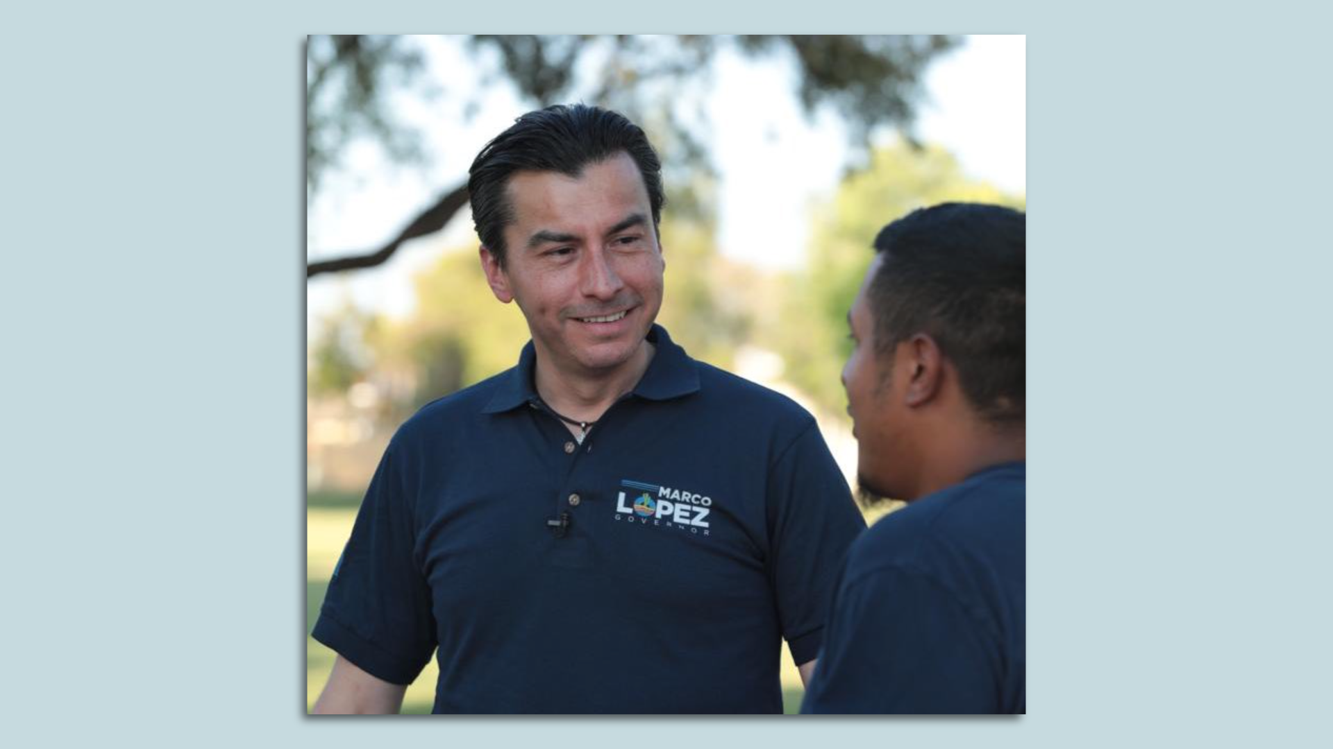 A man in a blue polo that says "Marco Lopez Governor" talking to another man.