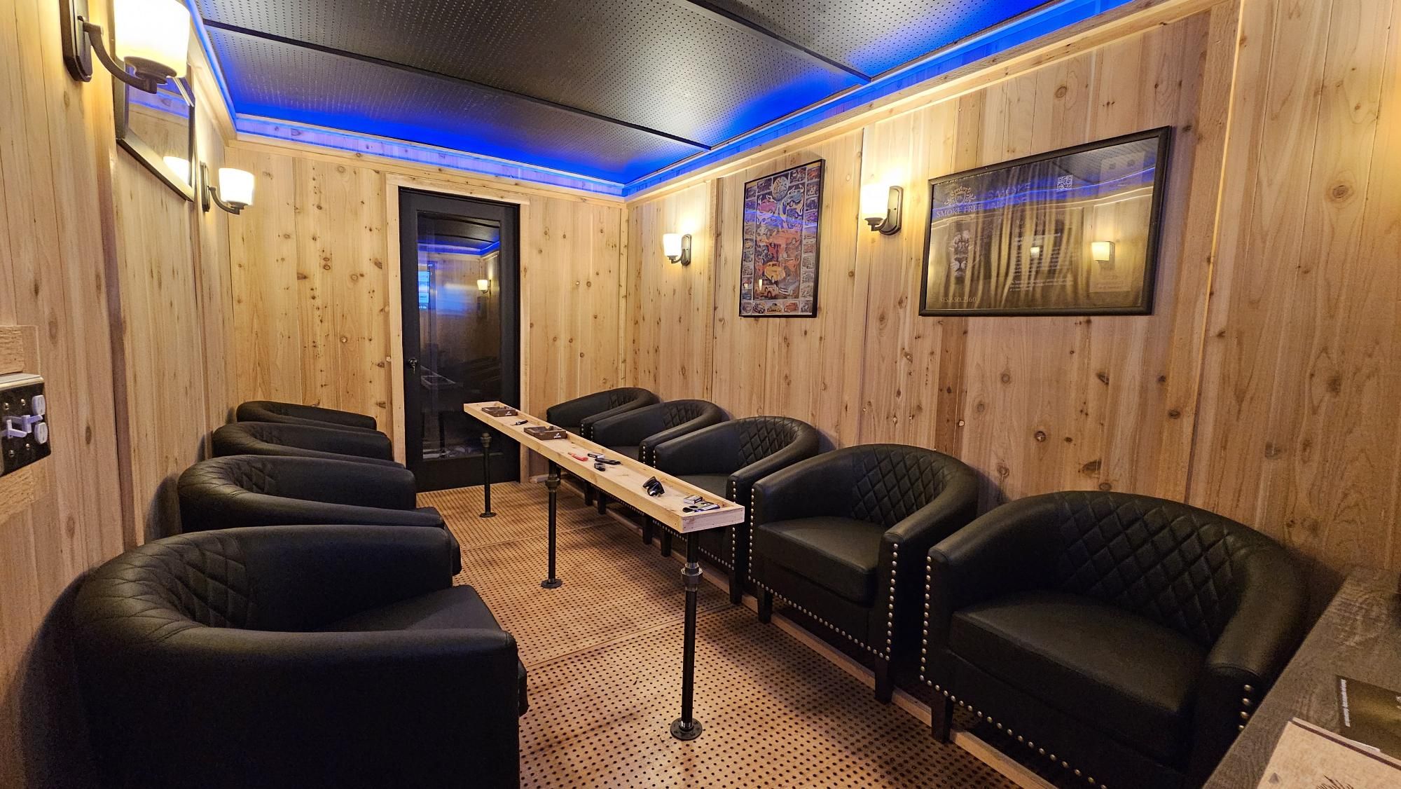 Wood-paneled lounge with a long light-wood table flanked by black tufted leather chairs. Blue ceiling lighting, wall sconces, framed posters, and a door at the far end.
