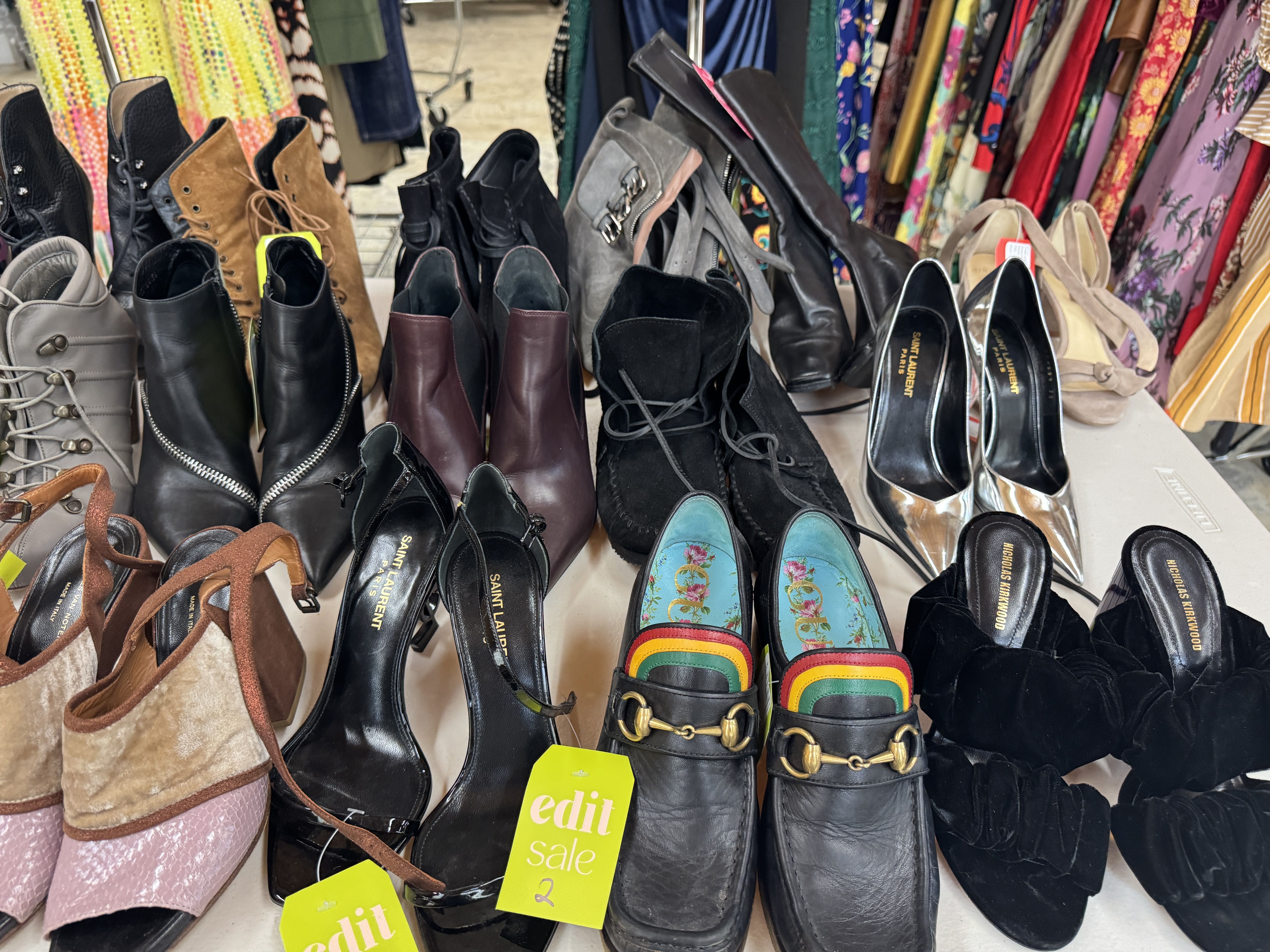 Various women's shoes displayed on a table, including boots, sandals, and heels in black, brown, metallic, and multicolor with sale tags, with colorful clothing racks in the background.