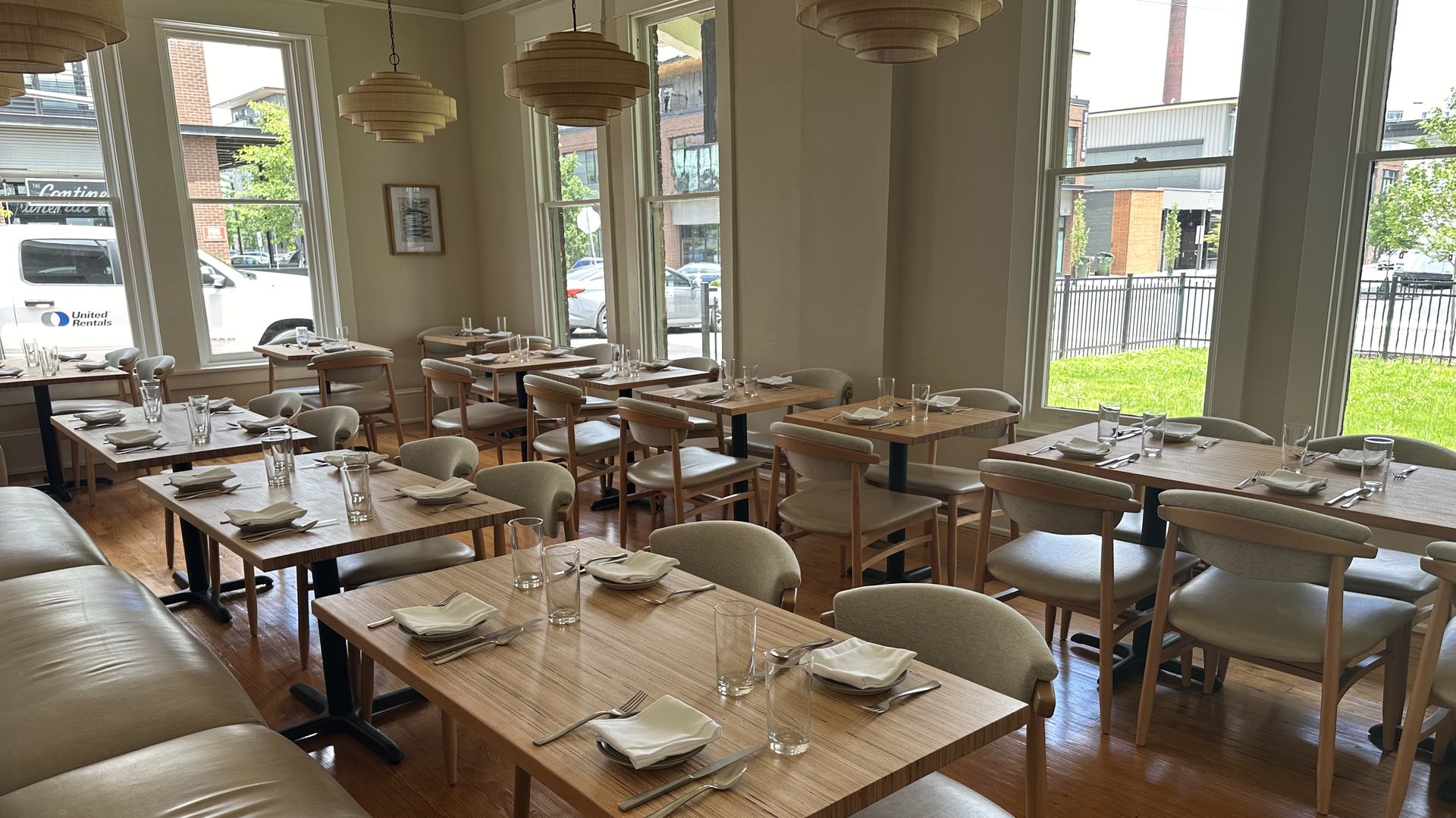 Bright cafe dining room with light wood tables and beige chairs, set with napkins, cutlery, and glasses. Large windows bring daylight; wooden pendant lamps hang from the ceiling; hardwood floors.