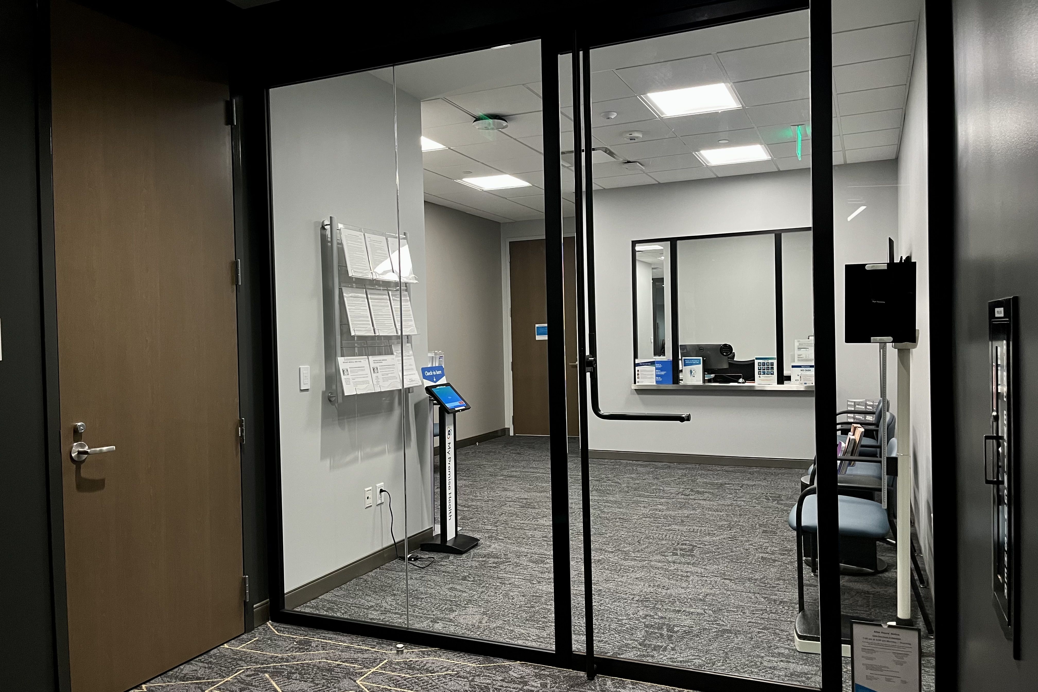 Empty modern waiting room with glass doors, gray carpet, blue chairs, a check-in kiosk, and a reception window with computer and signs behind it.
