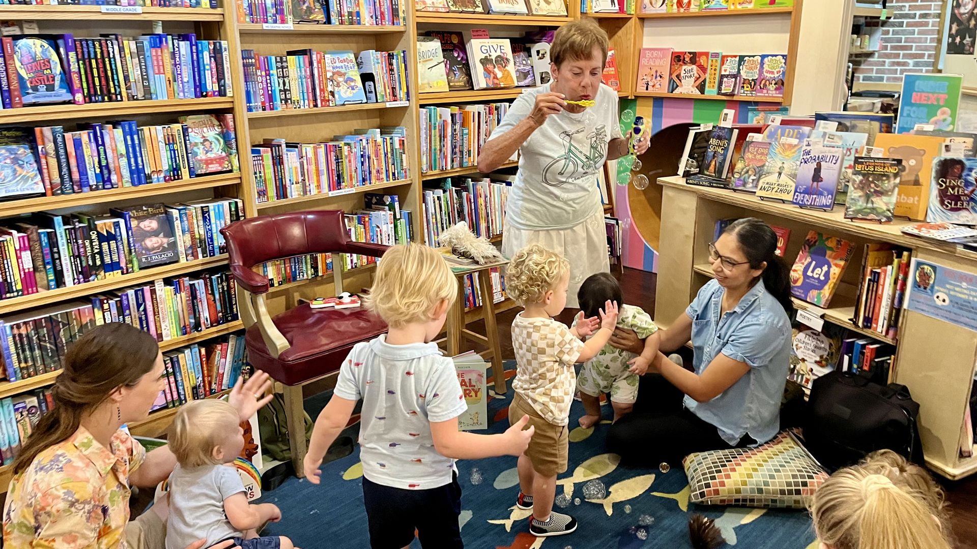 A woman, surrounded by shelves of colorful books, blows bubbles as children crowd around her.