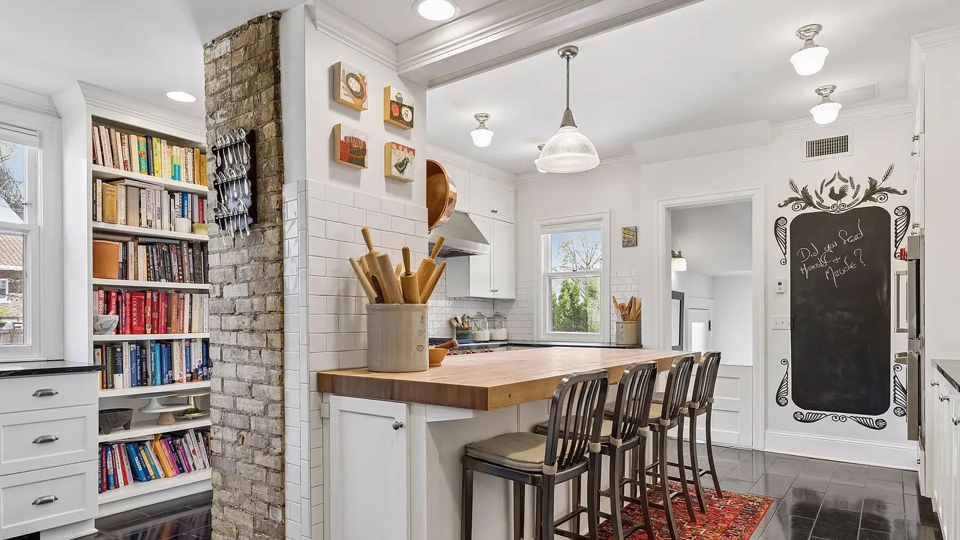 Bright kitchen with white cabinets, subway tile, and a wooden island with five metal stools. A brick pillar and bookcase wall separate the space; a chalkboard door with ornate trim and a red rug.