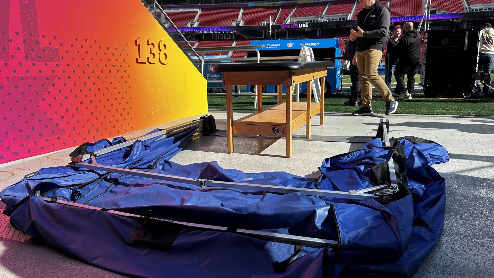 Blue collapsed tent and wooden table on concrete near stadium seats marked 138; people and NFL Super Bowl signage visible in background under bright sunlight.