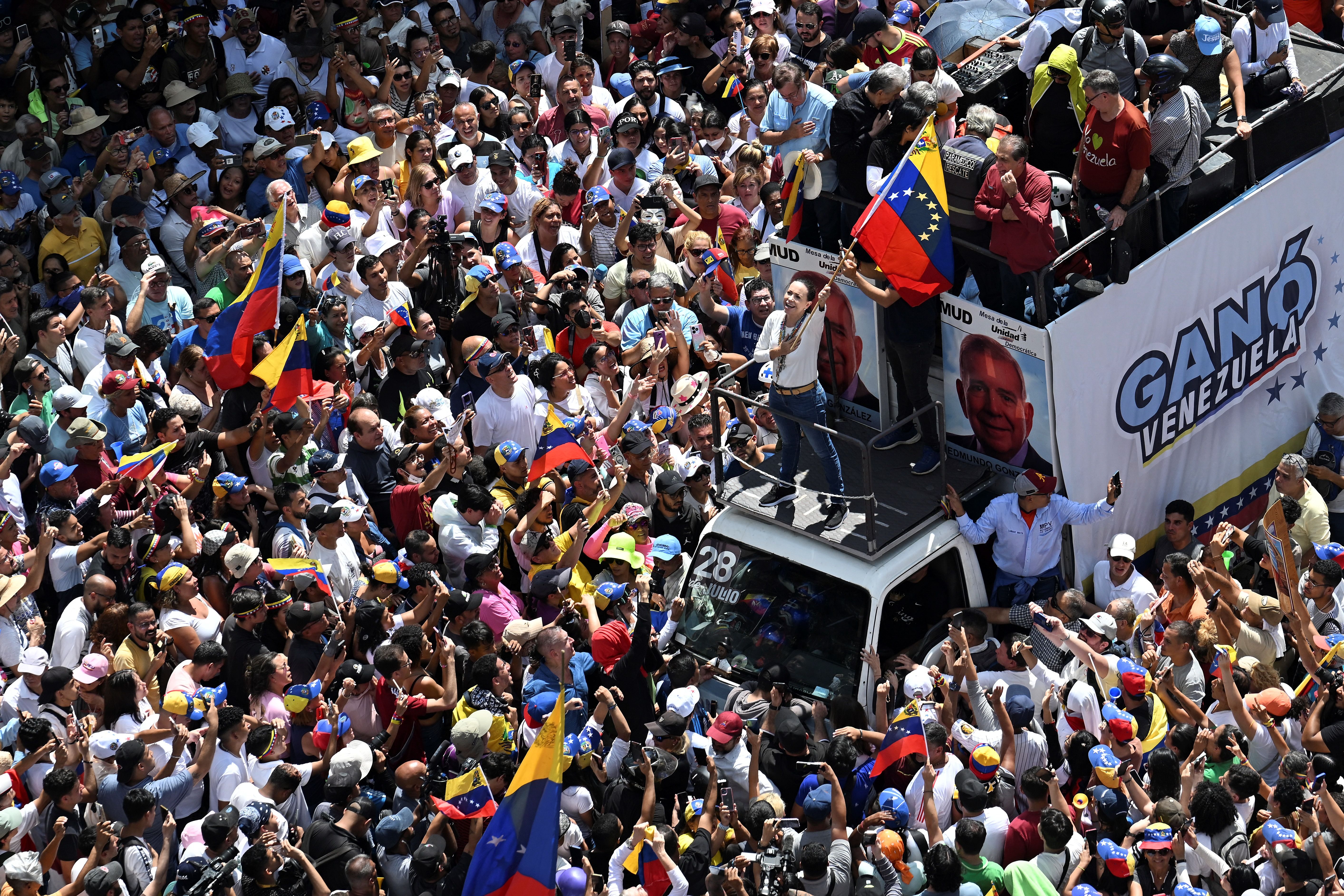 An aerial shot shows Maria Corina Machado waving a large Venezuelan truck atop a truck while surrounded by thousands of marchers 