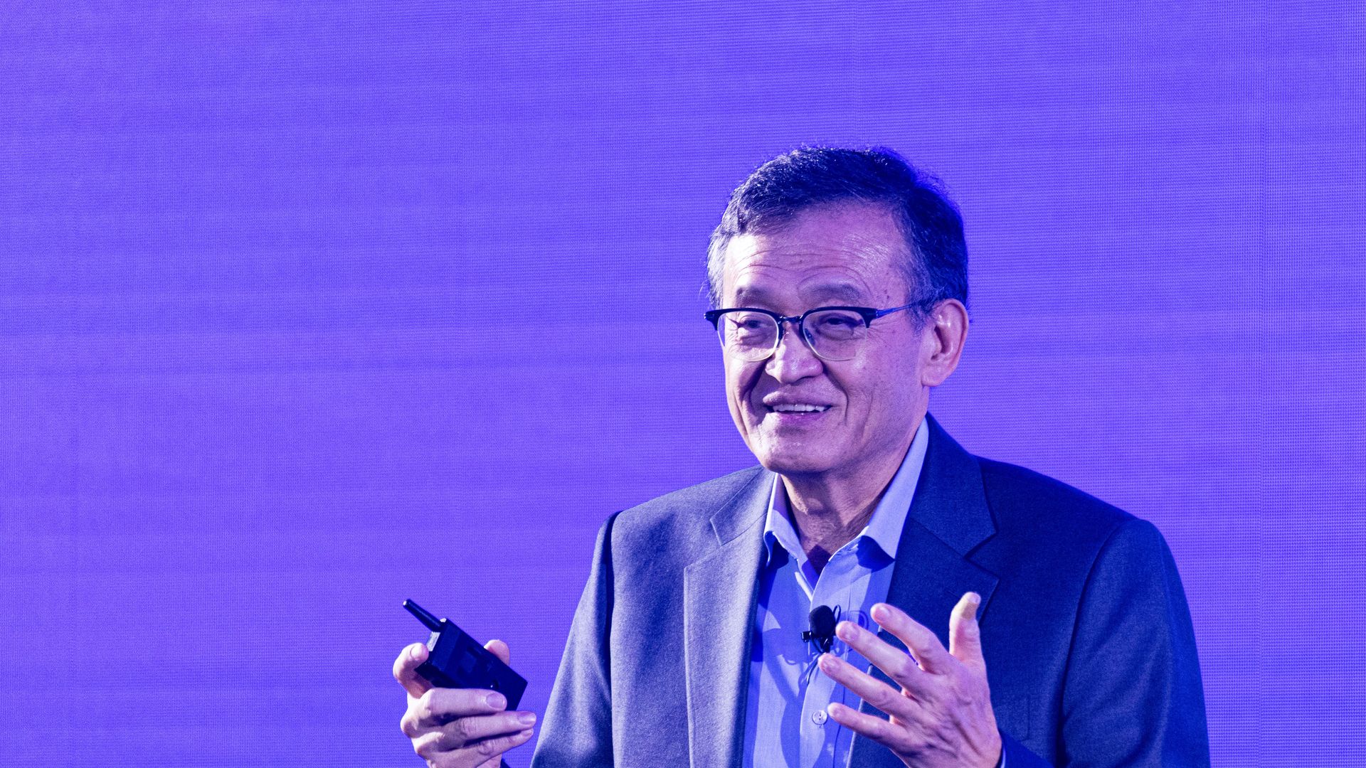 Man in glasses and a blue suit jacket speaking and gesturing while holding a device against a solid blue-purple background.