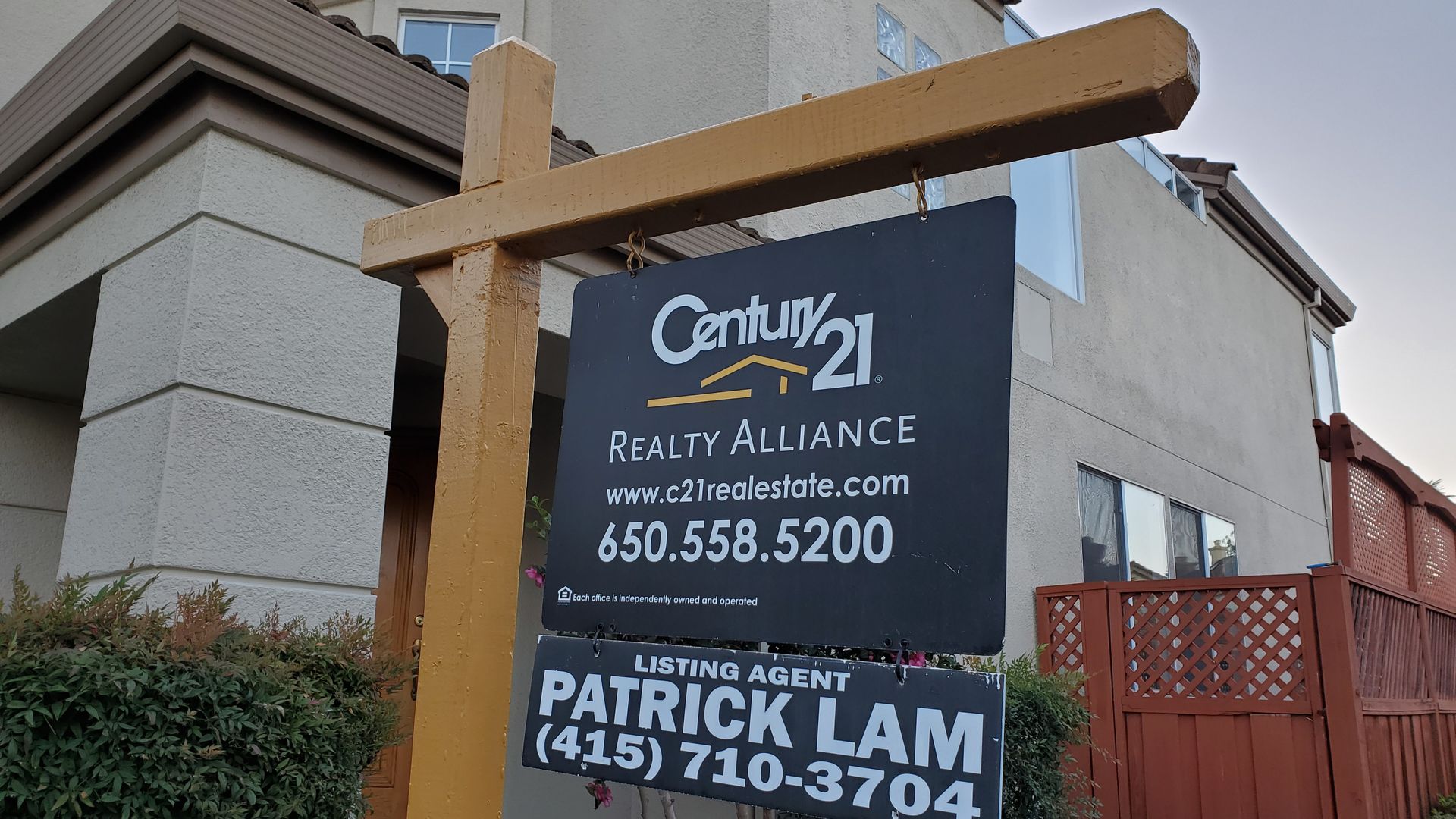 A black home-for-sale sign on a yellow post in front of a gray home with red fencing