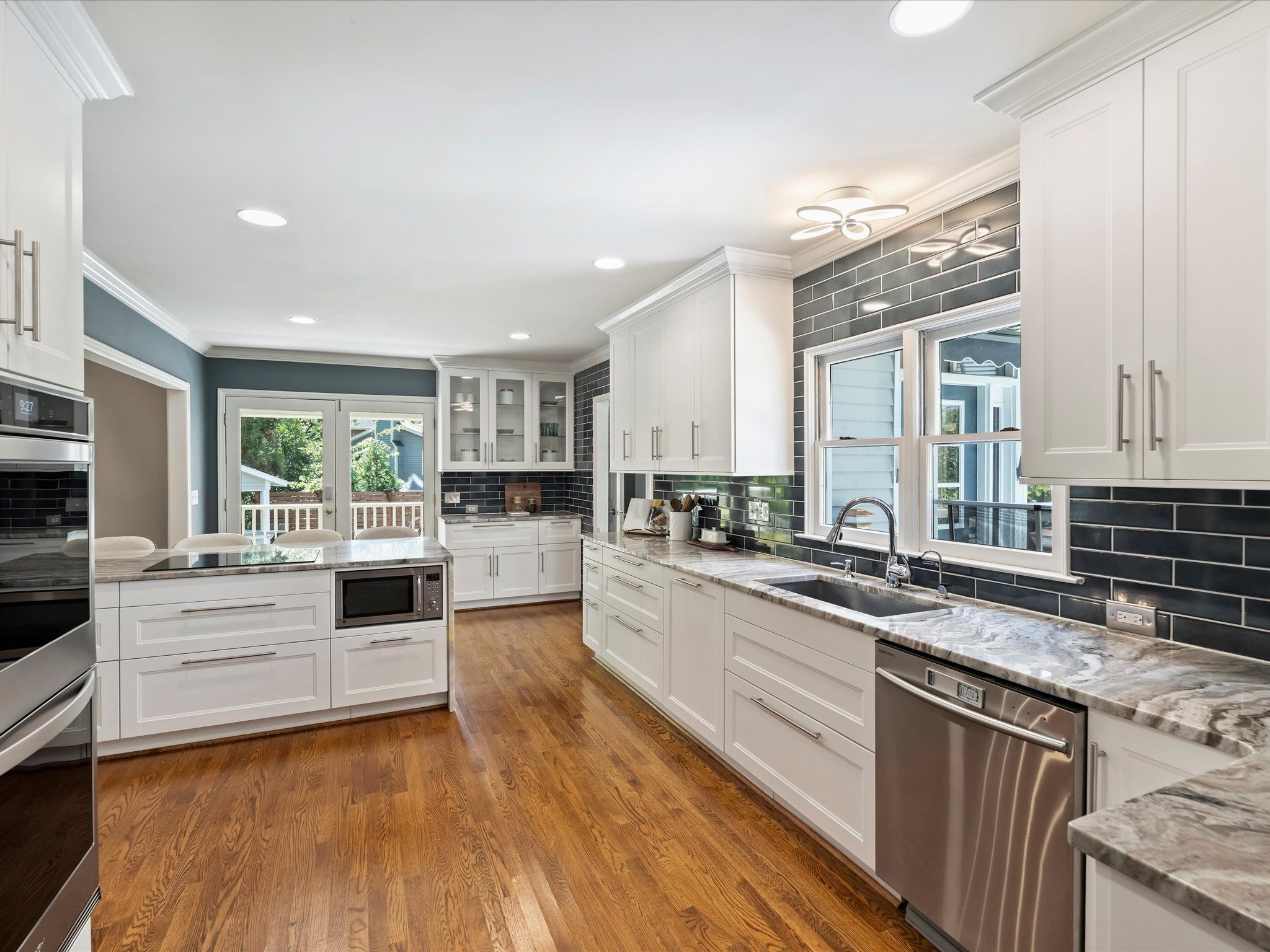 Bright modern kitchen with white cabinets, gray marble countertops, dark subway tile backsplash, stainless steel dishwasher, microwave, hardwood floor, and large windows with outdoor view.