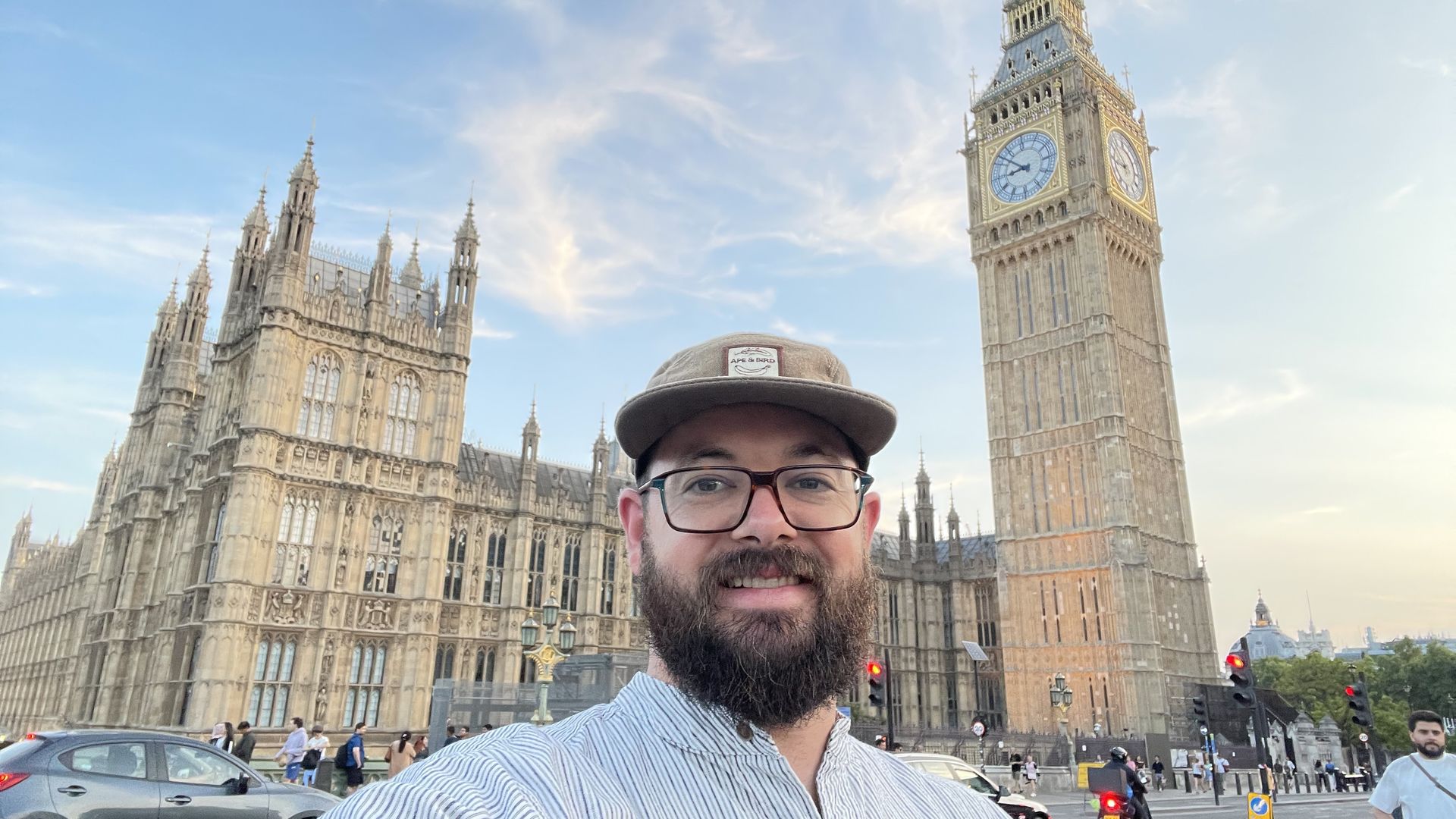 Selfie of a bearded man wearing glasses and a hat in the foreground, with London's Big Ben and the Houses of Parliament behind him on a clear day.