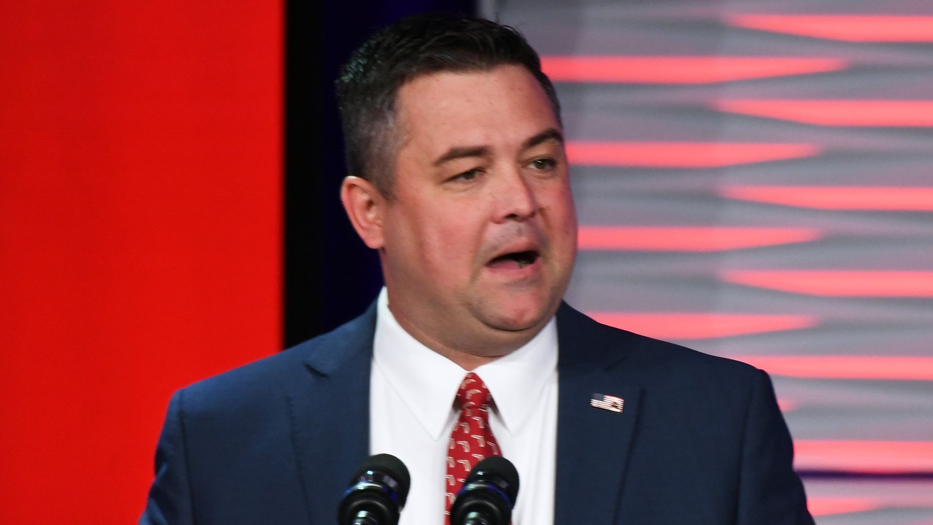A man in a navy suit, red tie and American flag pin speaking from a lectern.