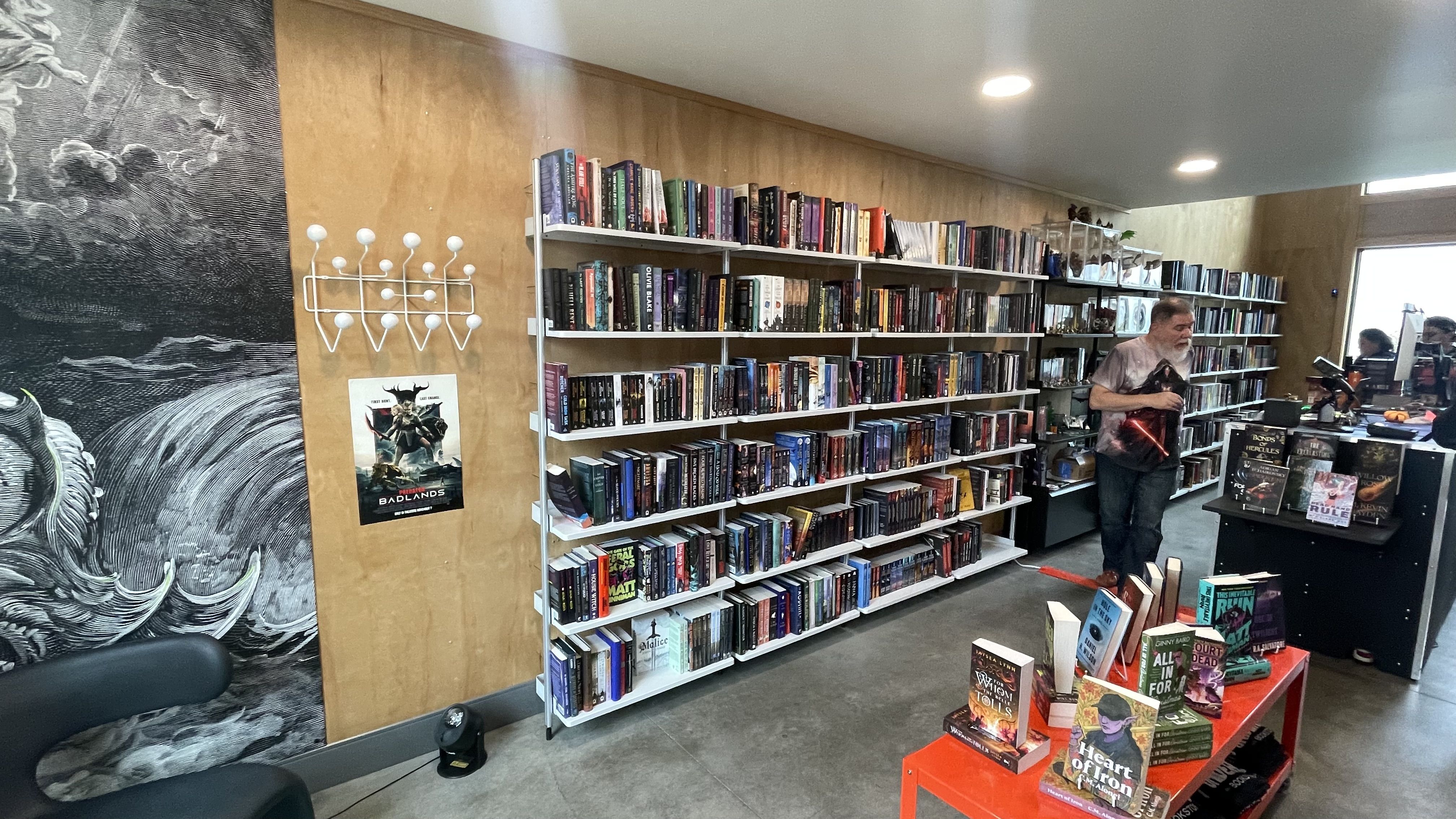 Interior of a bookstore with wooden walls, white shelves filled with books, a red table displaying books, a man standing near the shelves holding a book, and a black-and-white mural on the left wall.