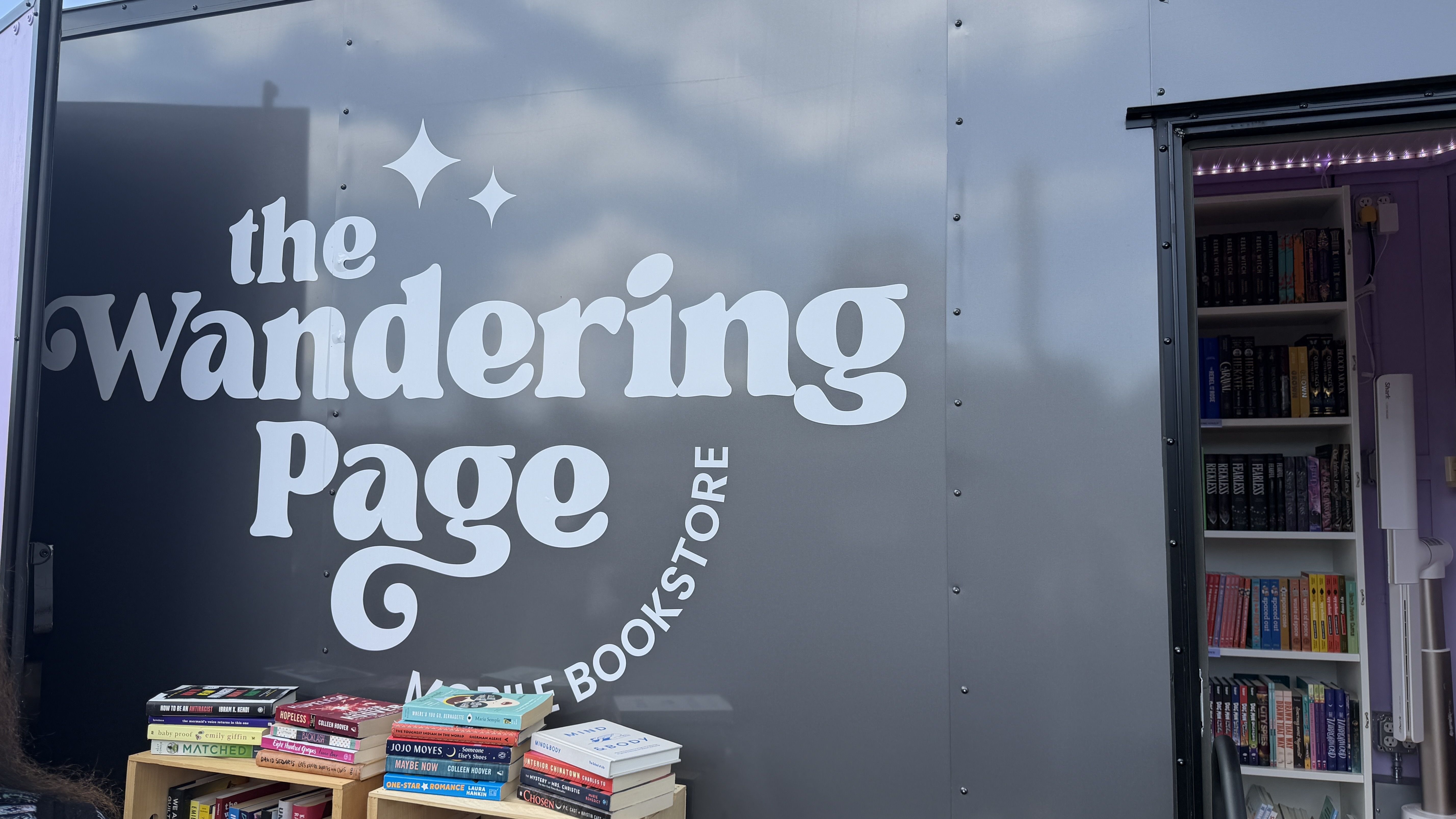 Gray mobile bookstore trailer with large white text the Wandering Page; open door reveals shelves of colorful books inside, purple interior and string lights, plus a table of stacked books outside.