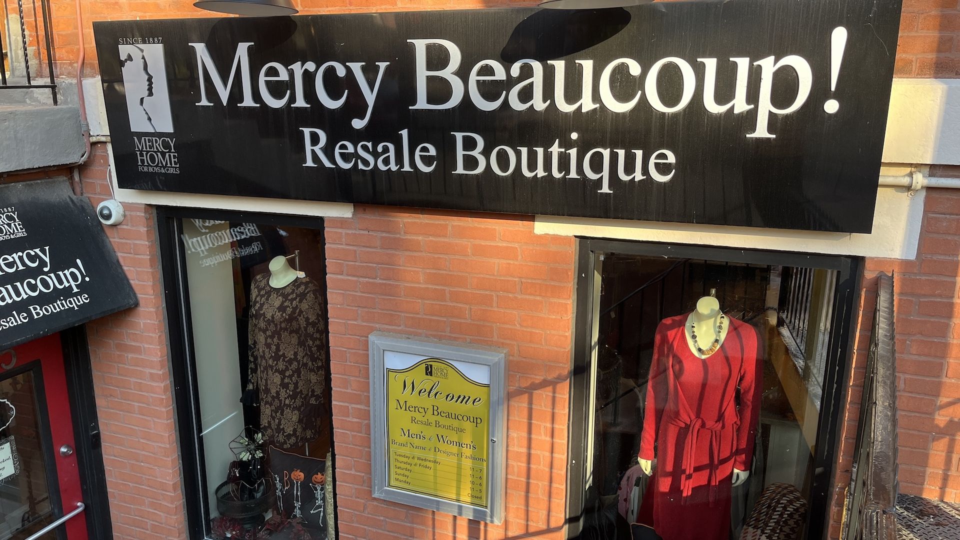 Black sign reading "Mercy Beaucoup! Resale Boutique" on red brick wall with two mannequins wearing dresses, one brown floral and one red with a necklace, and a yellow welcome sign below.