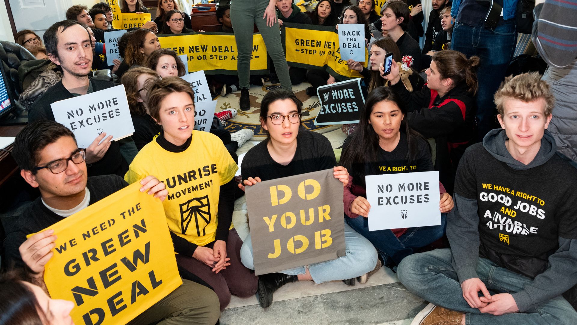 Youth activists sit with signs that read "green new deal" and "do your job"