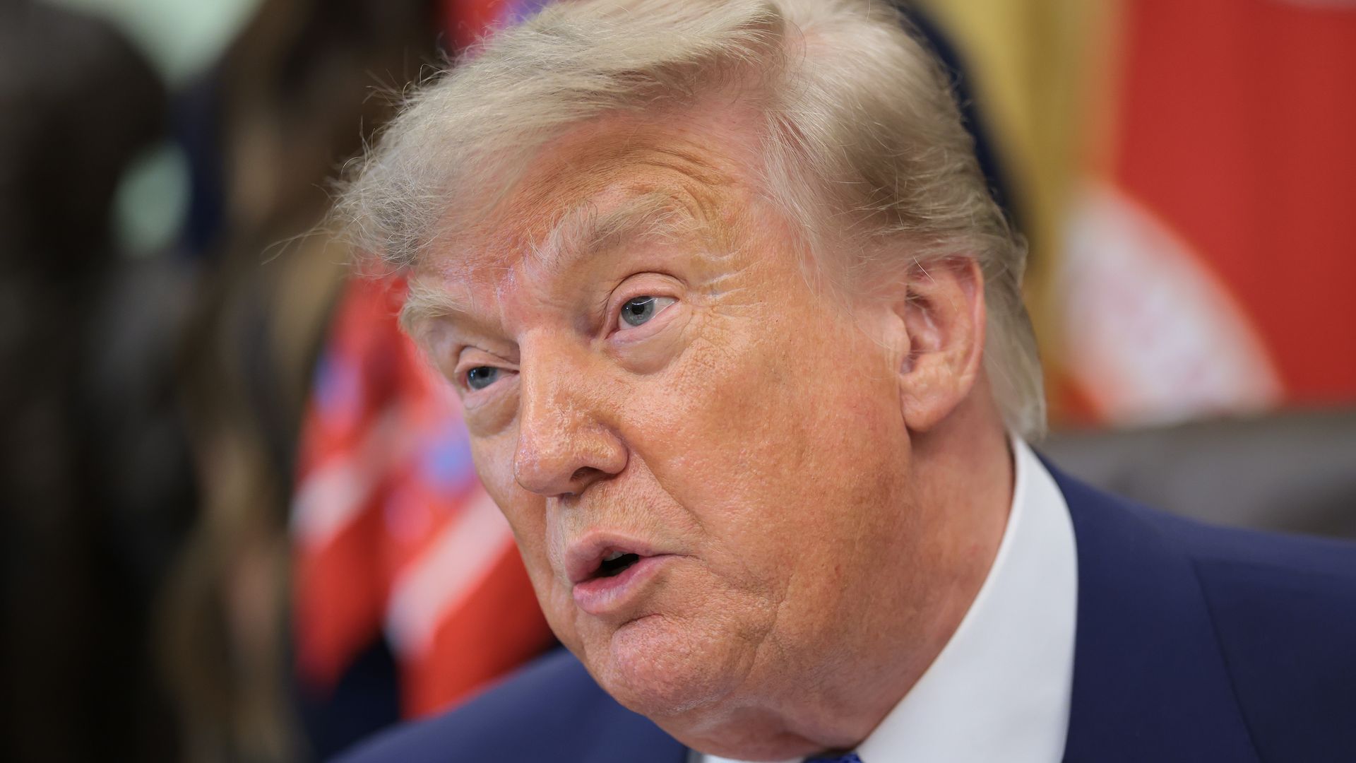 Close-up of a man with light hair in a blue suit and blue tie, mouth open as if speaking. Blurred red, white, and blue background suggests a patriotic event.