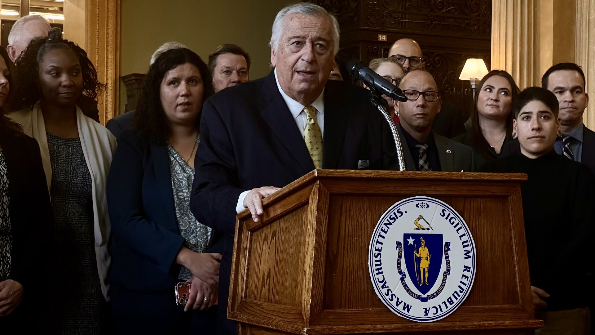 Mass. House Speaker Ron Mariano speaks from a lectern in the State House Library, surrounded by legislators and immigrant rights advocates on March 19, 2026.