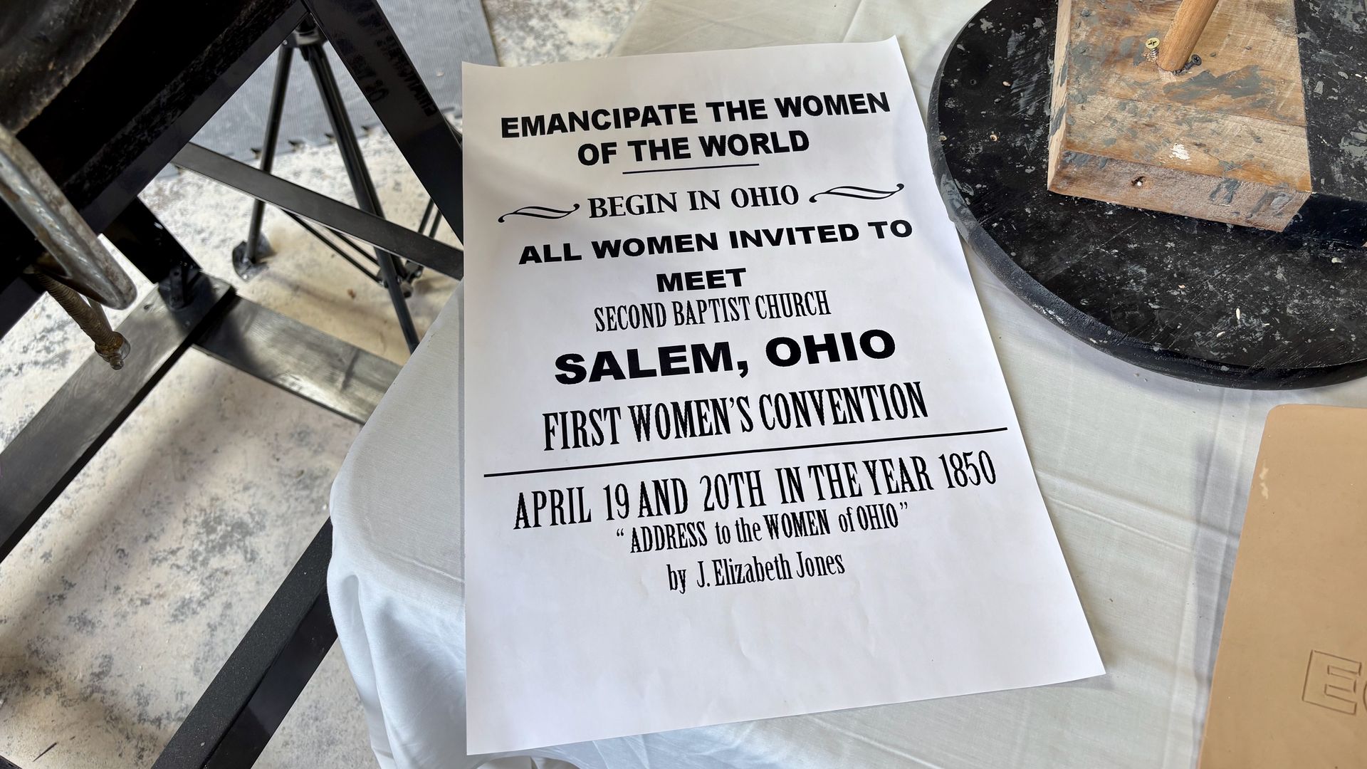 White poster on a worktable promoting the 1850 First Women’s Convention in Salem, Ohio. Bold lines read "Emancipate the Women of the World" and "Address to the Women of Ohio" by J. Elizabeth Jones.