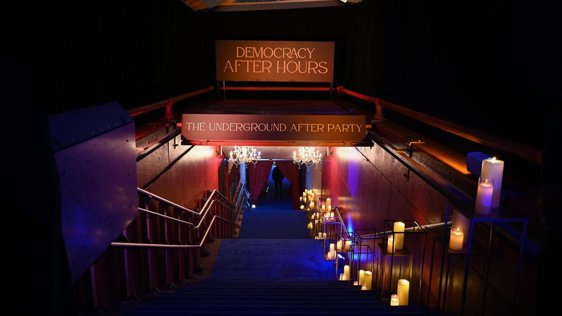 Dim venue stairwell descends into a club with warm amber lighting; signs read "DEMOCRACY AFTER HOURS" above and "THE UNDERGROUND AFTER PARTY" below, flanked by candles and chandeliers along the rails.