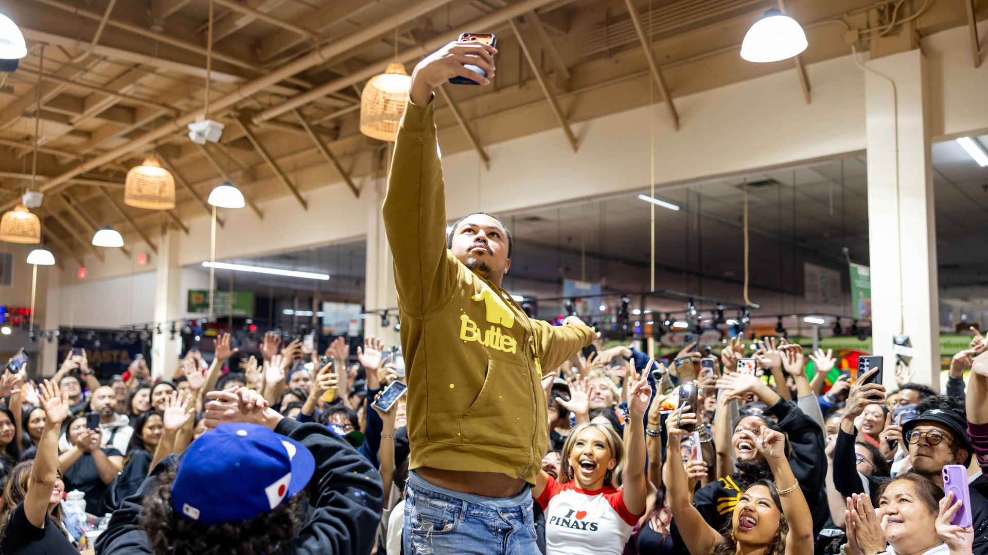 A man in a mustard-colored sweatshirt reaches up high to take a selfie among a huge crowd of people.