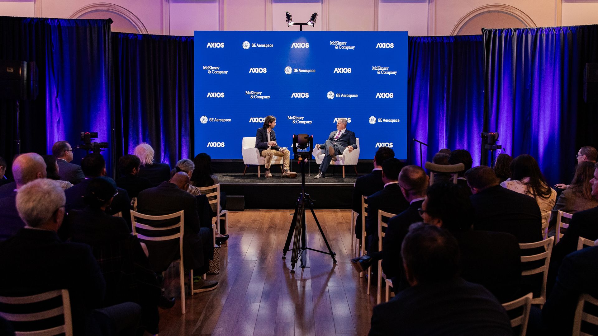 Two men sit on stage at a defense conference. The curtains flanking them are dark blue. The audience is in the foreground, darkened.