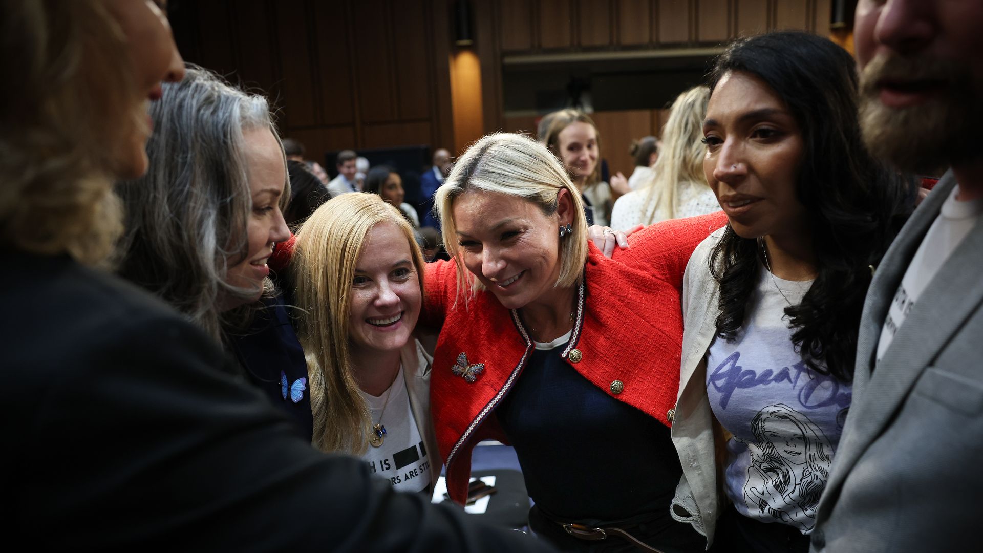 A woman in a red jacket, Dani Bensky, wraps her arms around a group of other women and one man after testifying on Capitol Hill. The people are emotional and smiling.