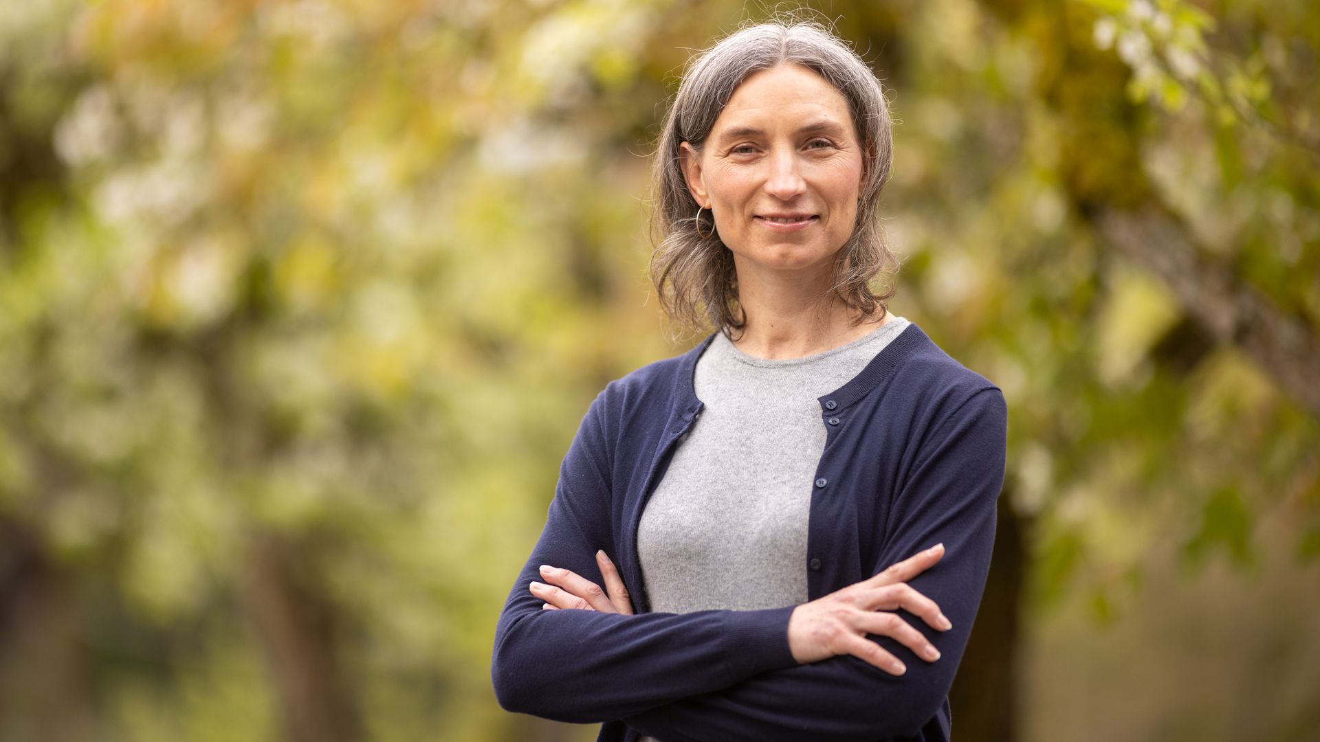 A woman in a gray shirt and blue sweater poses for a portrait with trees out of focus in the background.