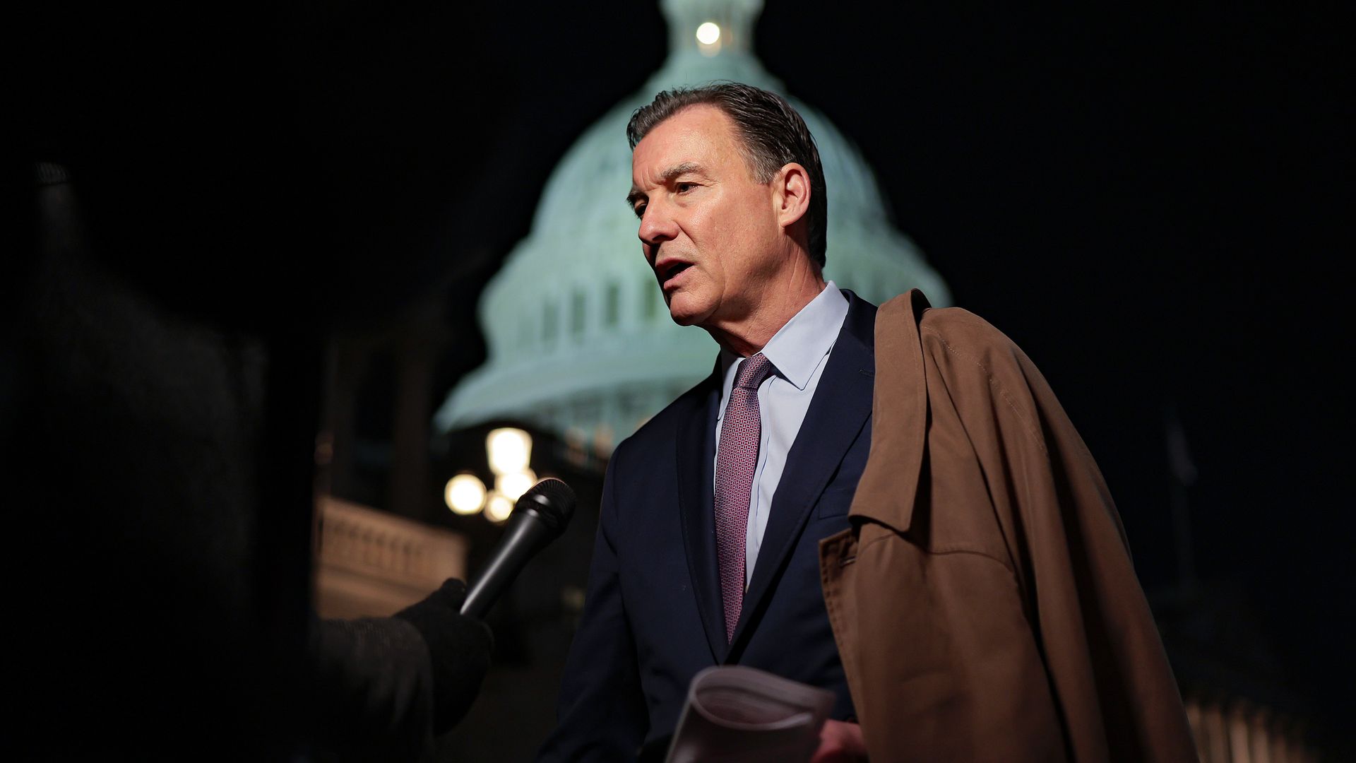 Man in a suit and tie speaking into a microphone outdoors at night, holding papers with a brown coat draped over his shoulder, Capitol building dome illuminated in the background.