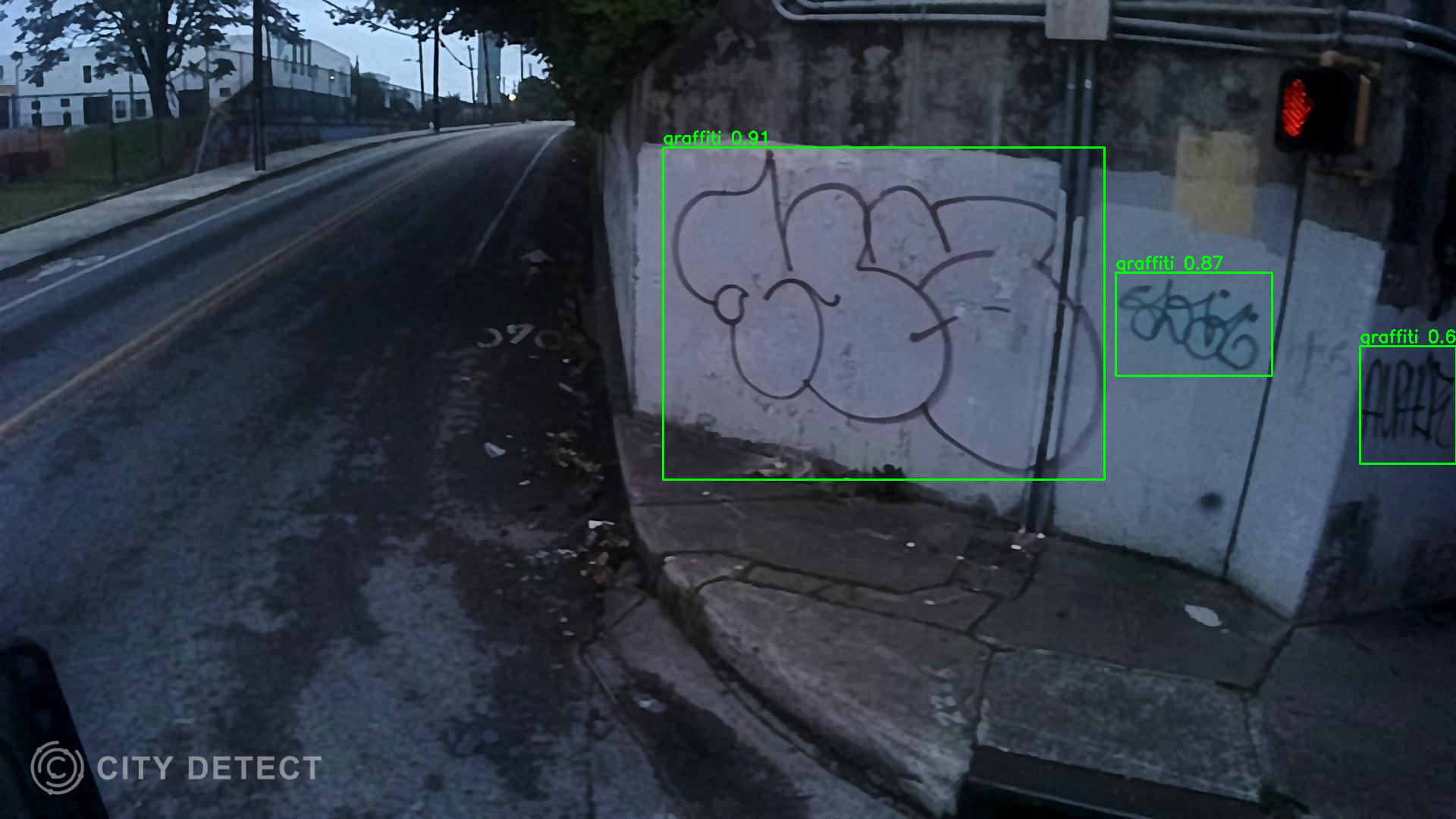 Street corner at dusk showing a concrete wall with black graffiti outlines and a red pedestrian signal. The road curves uphill with trees and buildings in the background.