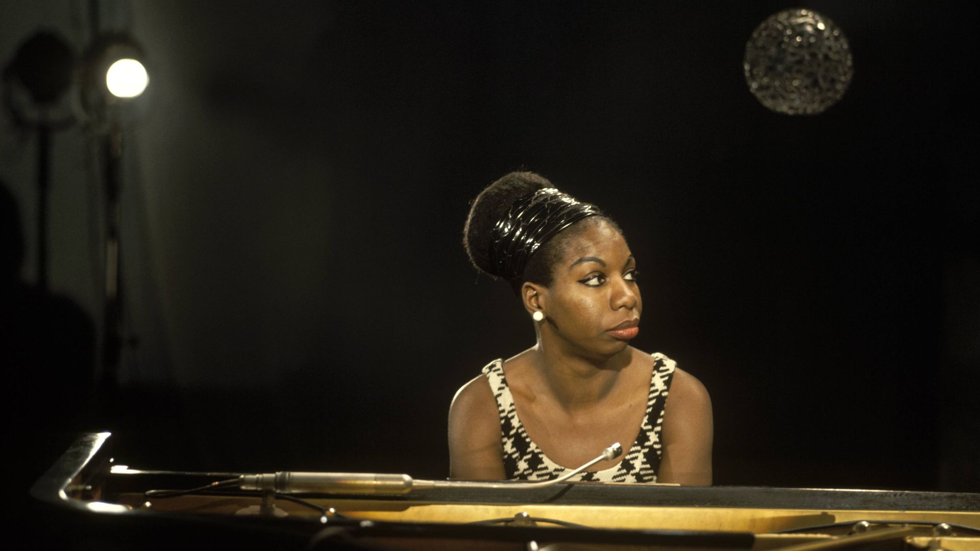 Nina Simone with a patterned sleeveless top and an updo hairstyle sits at a grand piano, looking to the side in a dimly lit room with hanging spherical lights and a spotlight in the background.