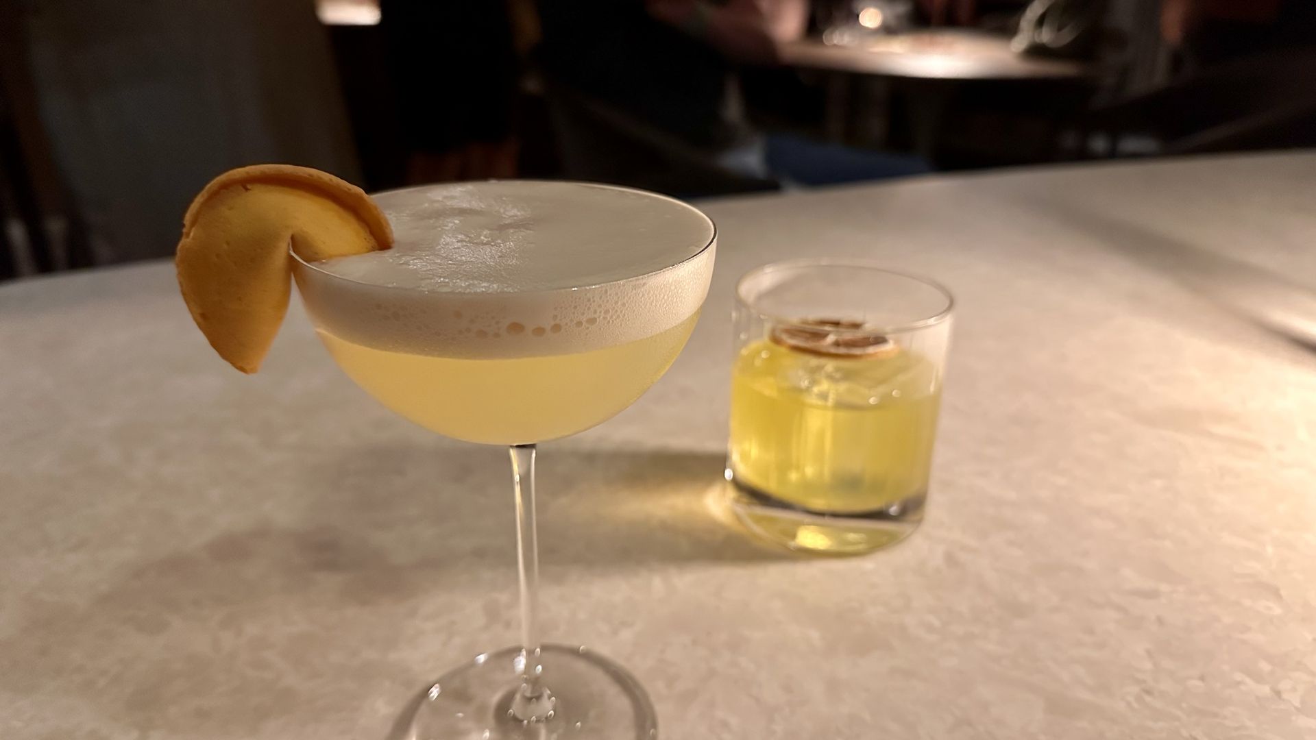 Pale yellow foamy cocktail in a coupe with a lemon twist garnish in the foreground; a short glass with lemon slices sits in the background. Warm bar lighting.