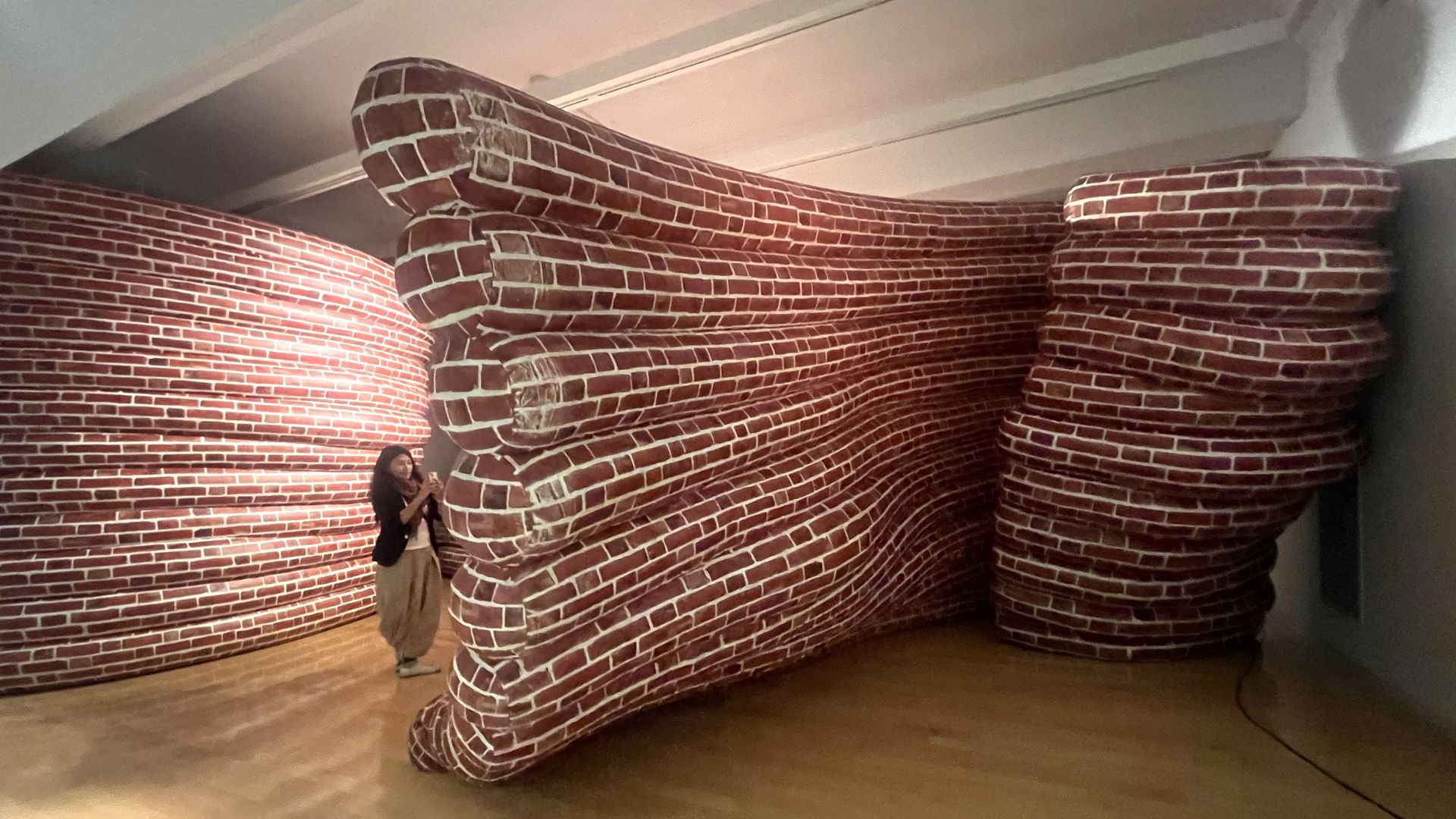 Woman in beige pants and black jacket stands next to large, curved sculpture painted to resemble red brickwork with white mortar lines in a gallery space.