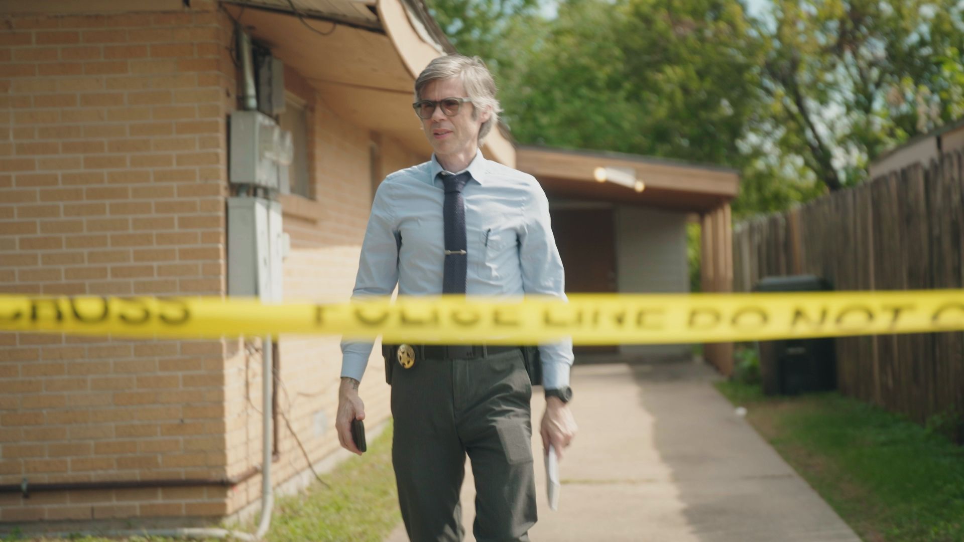 A man in a light blue shirt and tie walks through a police scene, indicated by yellow police tape in the foreground. He is holding a notebook and appears to be involved in an investigation. The setting is outdoors, with a brick building on the left and a wooden fence on the right.