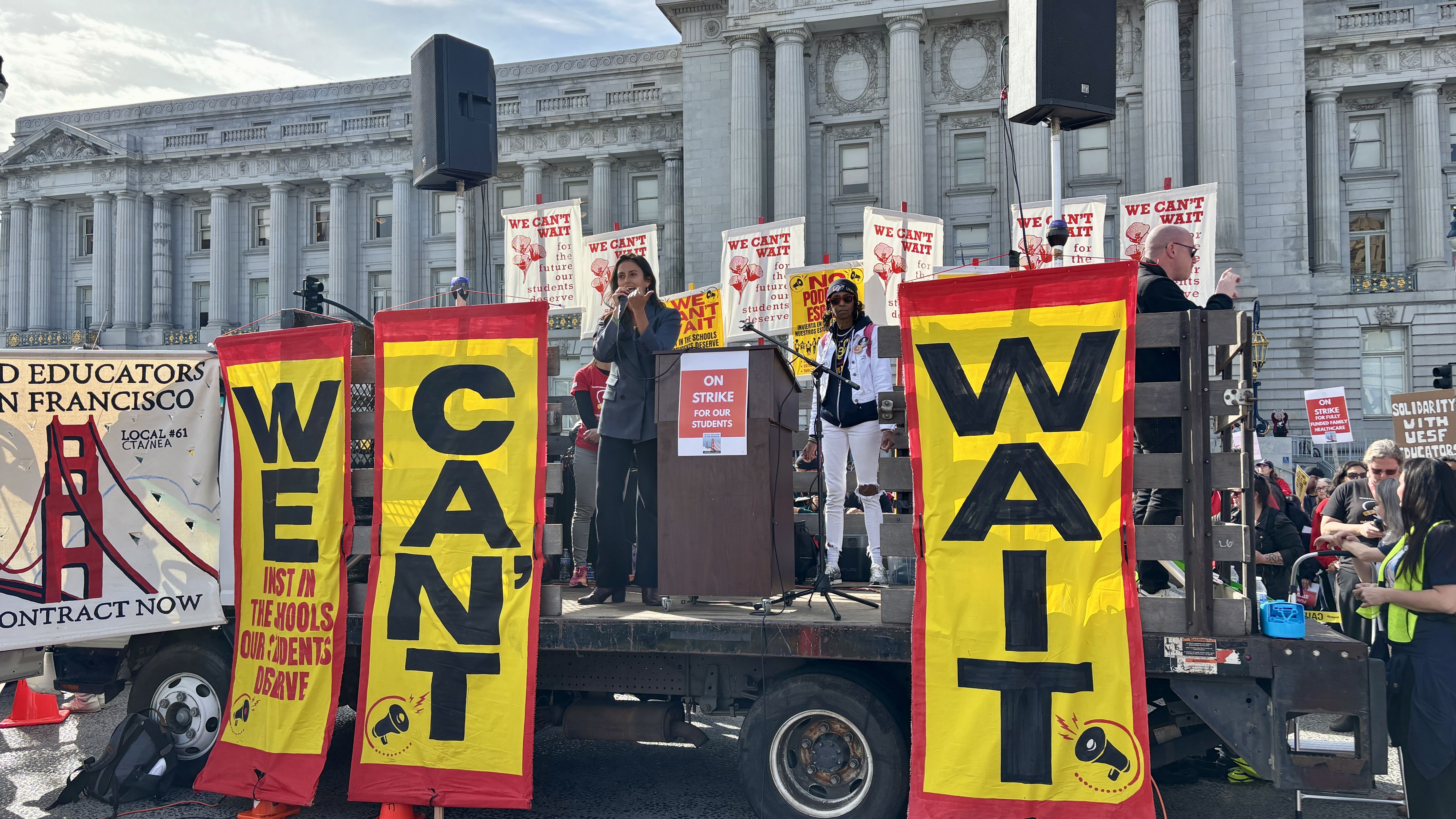 People at a strike rally in front of a large classical building hold yellow and red banners spelling "WE CAN'T WAIT" and signs supporting educators and students in San Francisco.