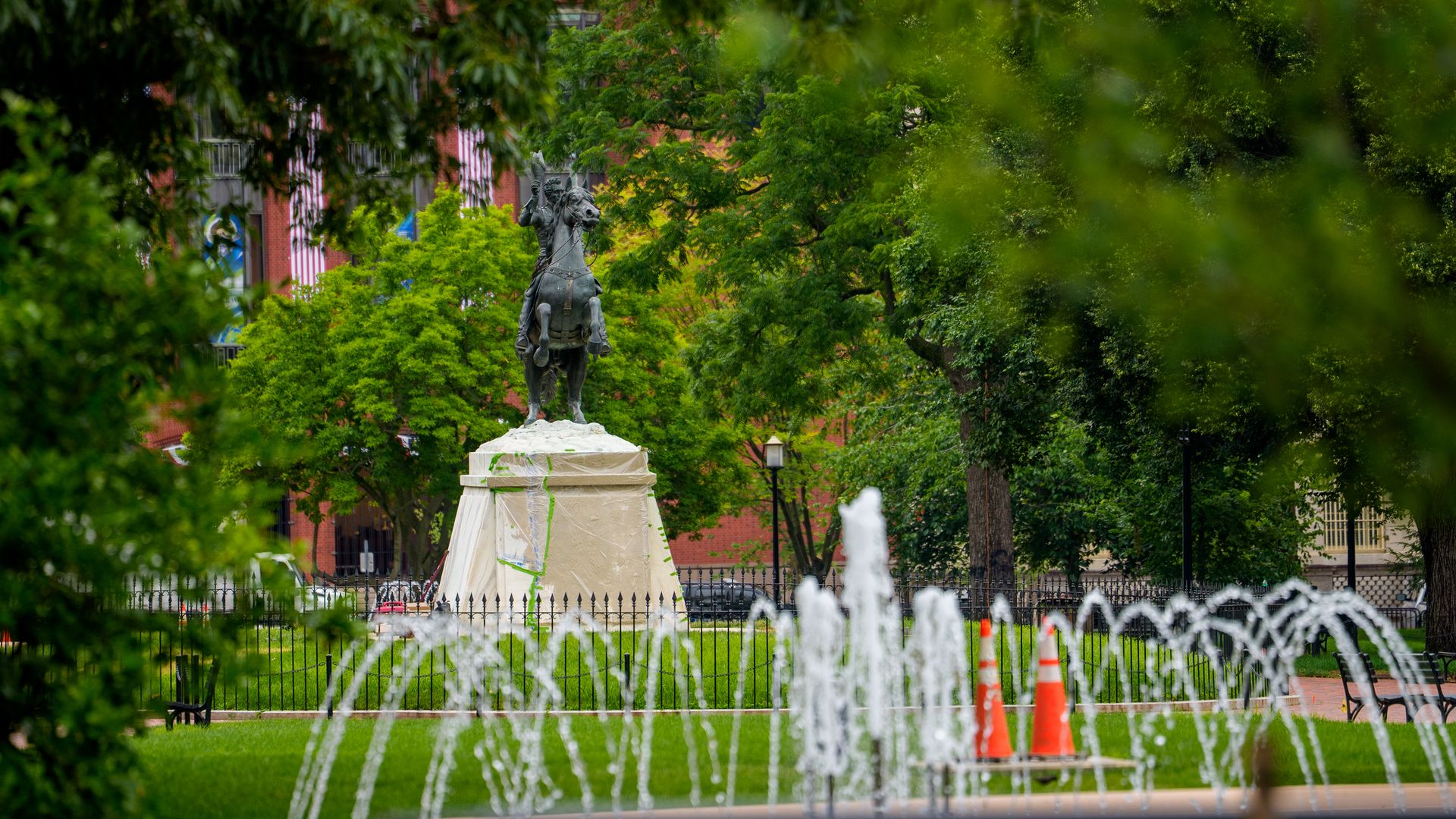 Bronze statue of a mounted rider on a pedestal in a city park, framed by dense green trees. Fountains shoot water in the foreground, with a red brick building visible behind.