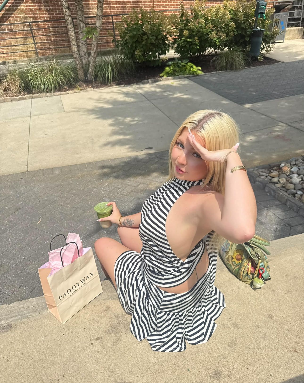 Blonde woman in a black-and-white striped halter dress kneels on a sidewalk, shielding her eyes from the sun with one hand, holding a green drink; shopping bags nearby, brick wall and greenery behind.