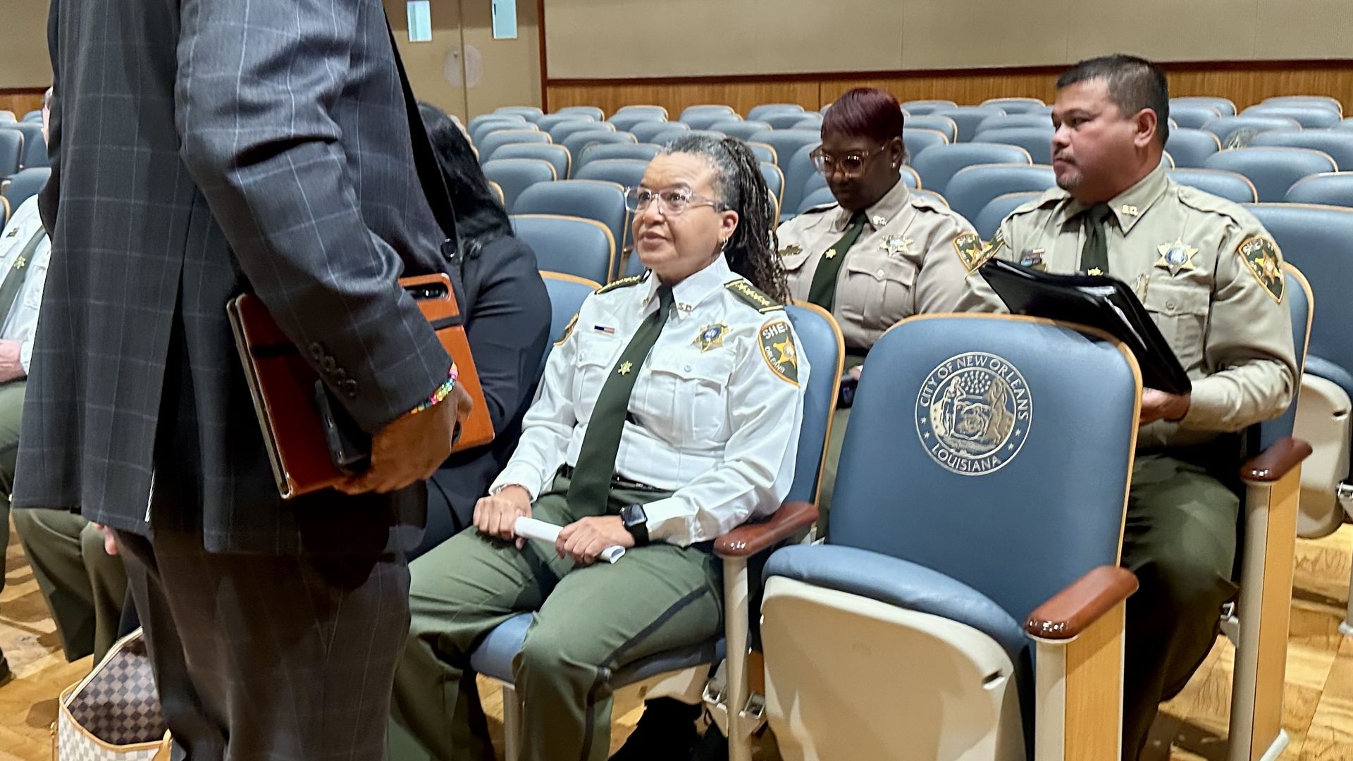 Auditorium with blue seats and several uniformed officers seated. A man in a dark suit stands at left holding a folder, while a woman in a light-blue sheriff uniform sits center.