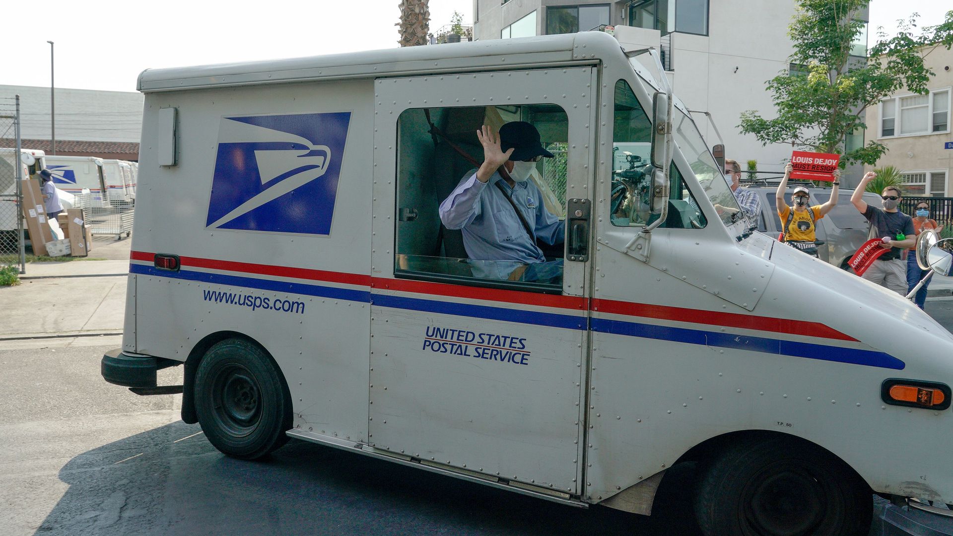 A mailperson drives a USPS truck past a crowd of people holding signs on the side of a road