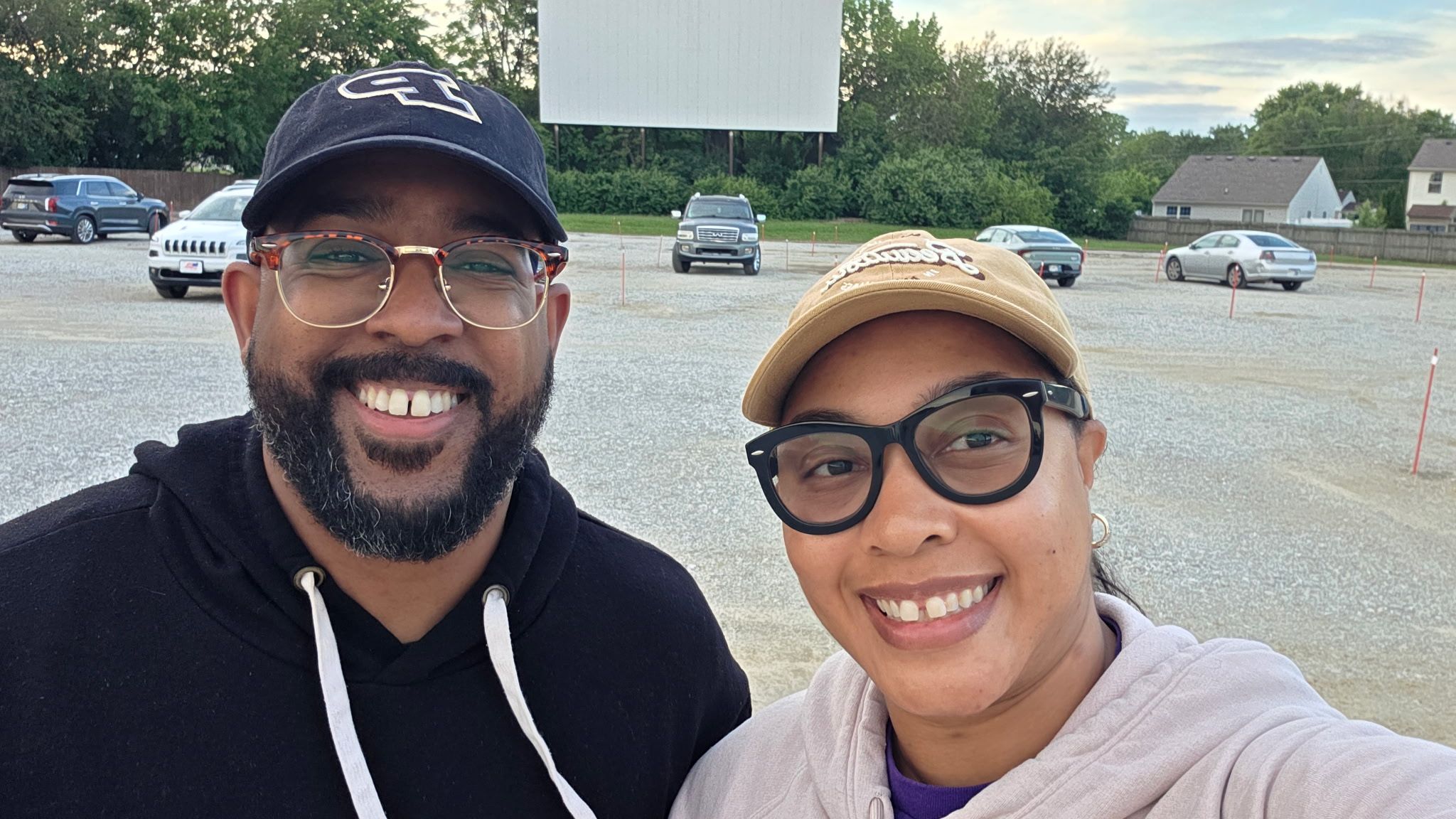 Two friends take a selfie in a gravel parking lot, smiling. The man wears a navy cap and glasses, the woman a tan cap and black glasses, with cars and a large blank billboard behind them.