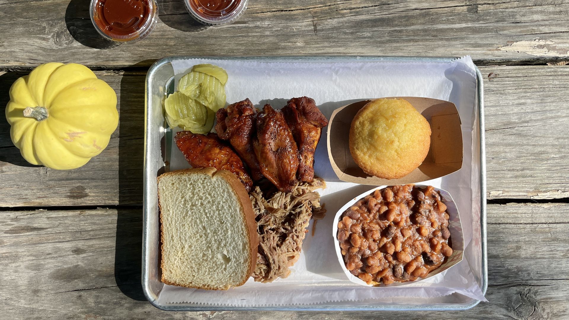 Barbecue meal on a tray with sliced white bread, pulled pork, glazed chicken wings, pickles, baked beans, and a cornbread muffin next to a yellow squash and two cups of BBQ sauce.