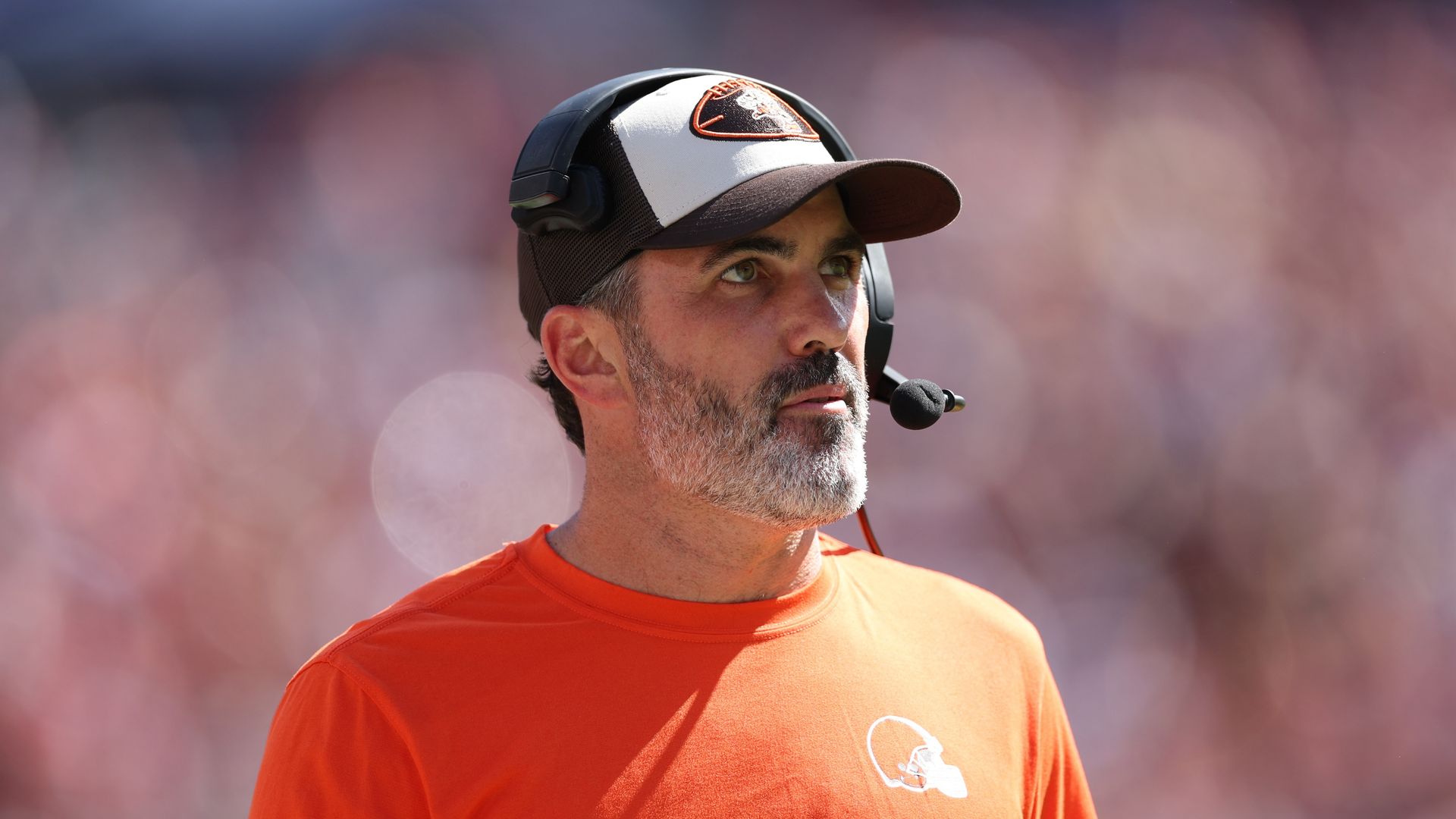 Browns quarterback Kevin Stefanski walking the sideline in an orange shirt, hat and headset. 
