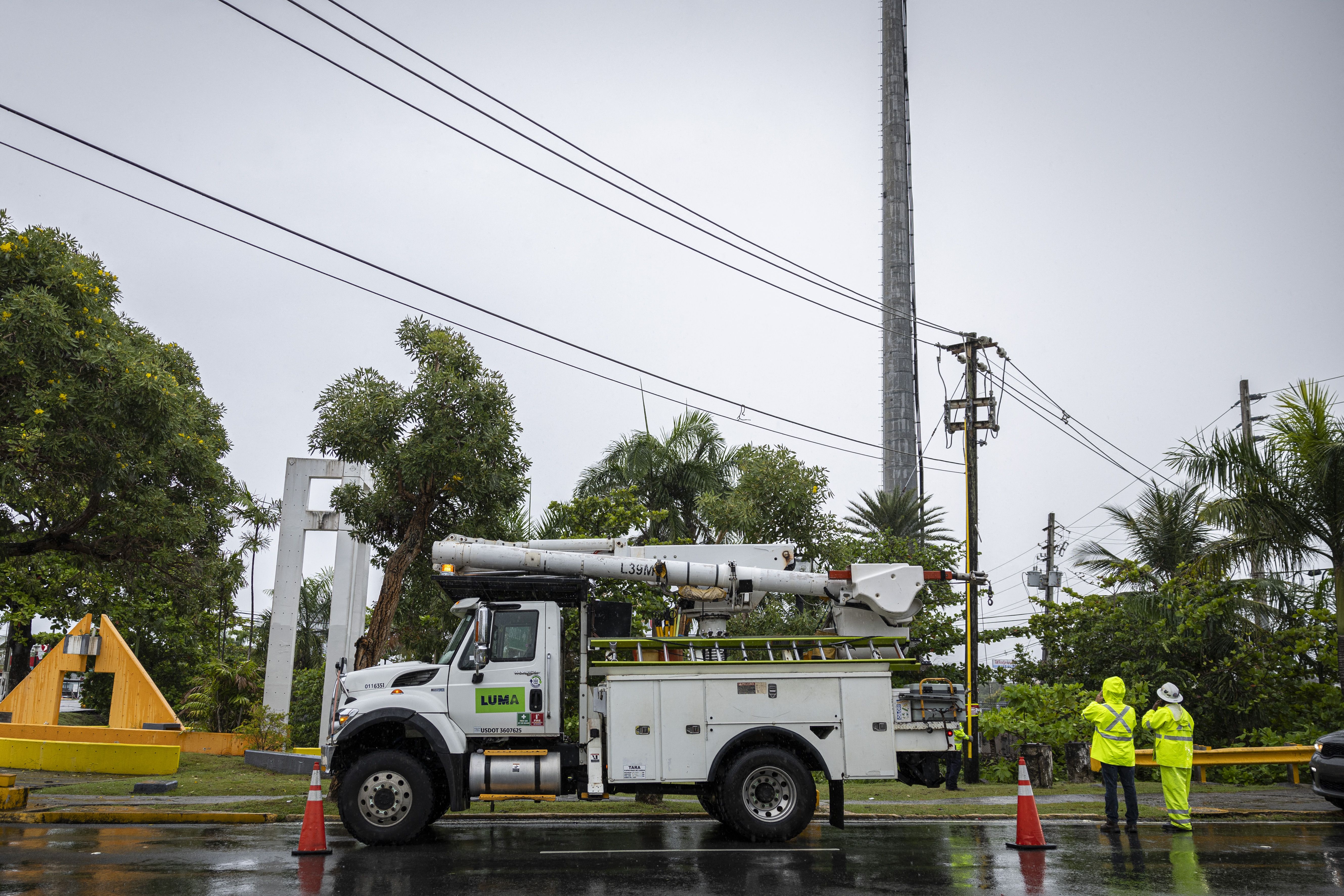 White utility truck with "LUMA" logo parked on wet road with orange cones, two workers in bright yellow rain gear talk near power lines on a cloudy day with green trees in background.