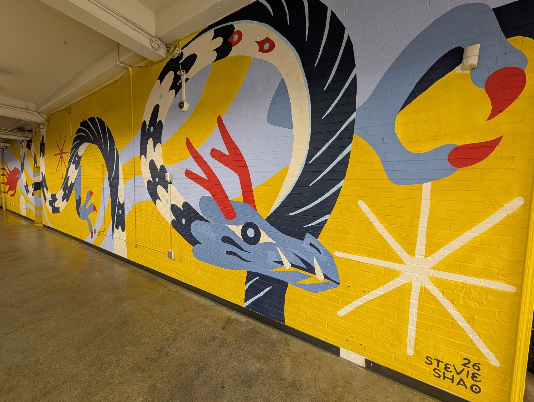 Indoor corridor featuring a bright yellow mural on a brick wall with abstract blue, black, red shapes and swirling patterns. Concrete floor, exposed ceiling beams, and a security camera with wires.