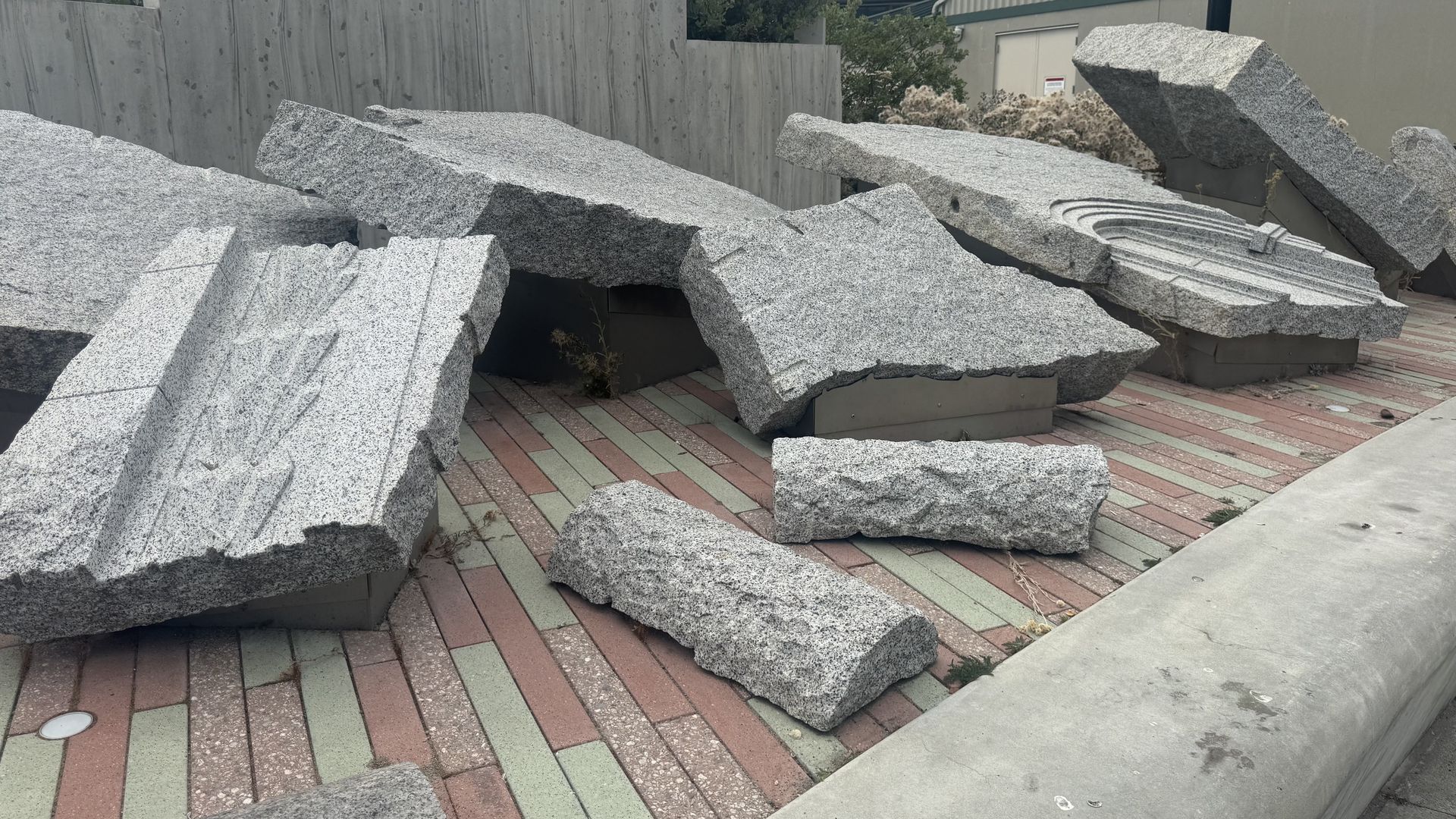 A pile of large gray granite rocks and broken architectural fragments scattered across a red-green tiled courtyard; a concrete wall and building in the background; carved grooves.