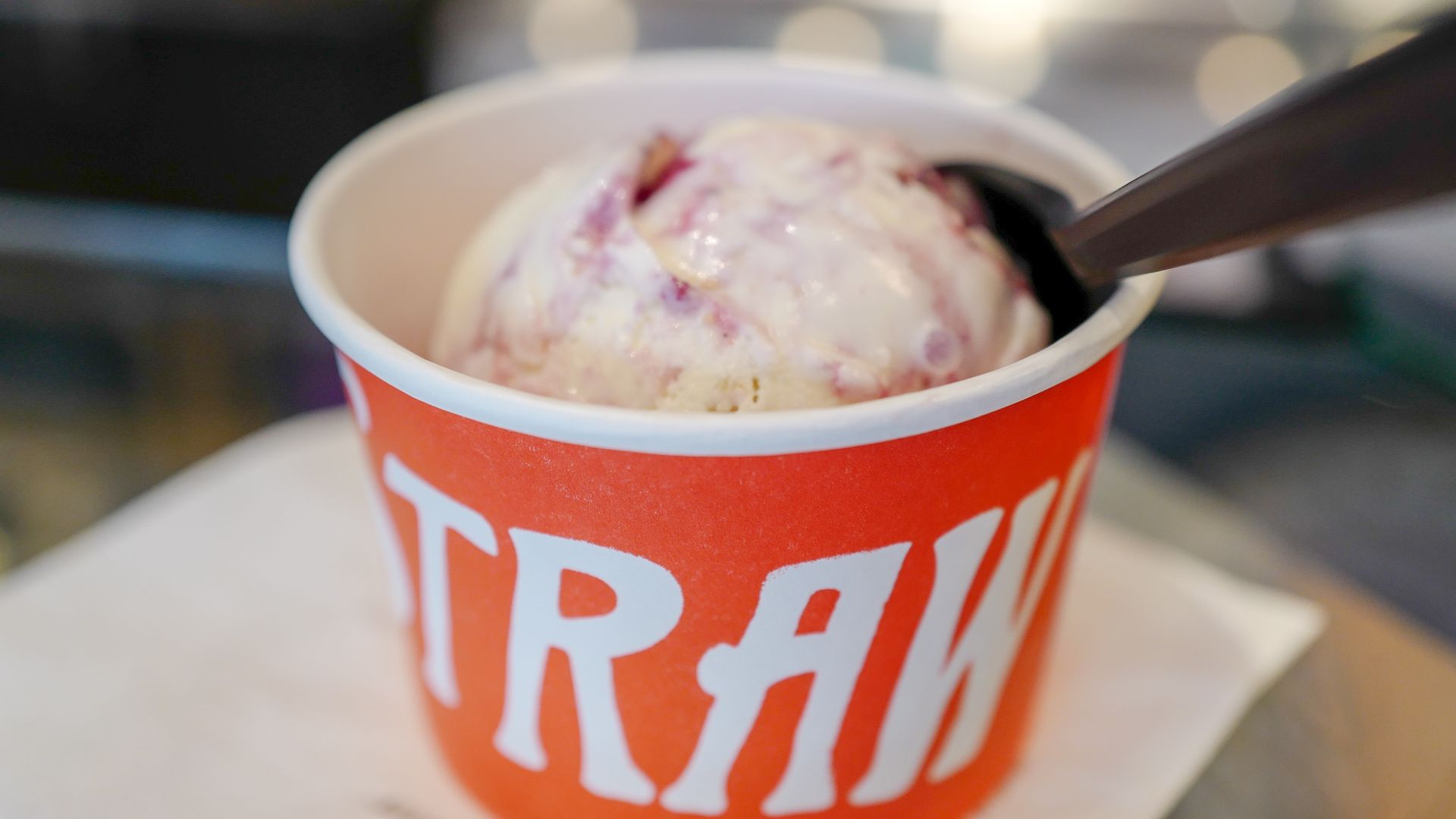 Close-up of a scoop of ice cream in a red cup with white lettering, a dark spoon resting on the rim; blurred background with warm bokeh lights.