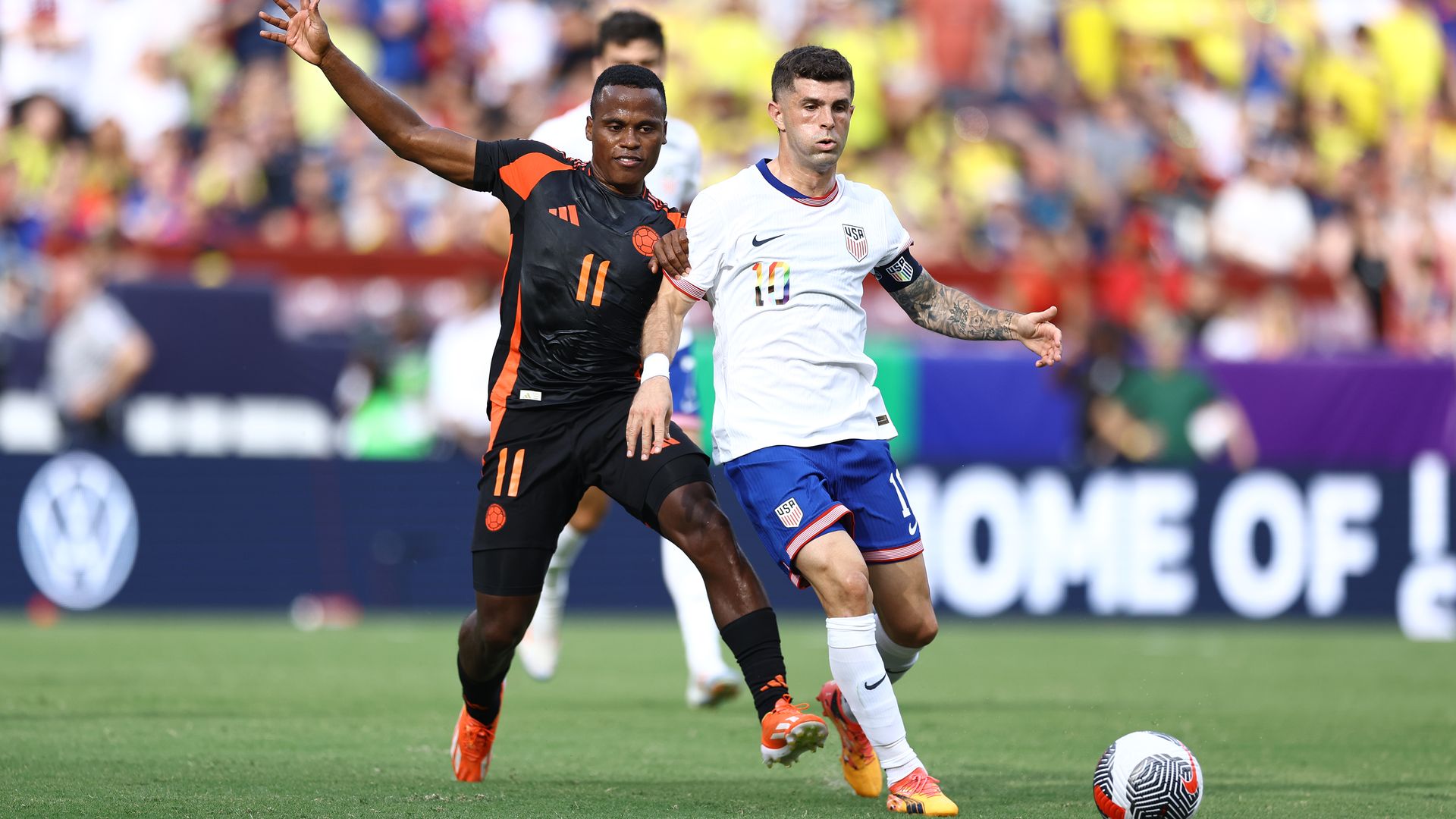 A soccer player in a white jersey runs toward a ball while another player in a black jersey follows closely behind him. 
