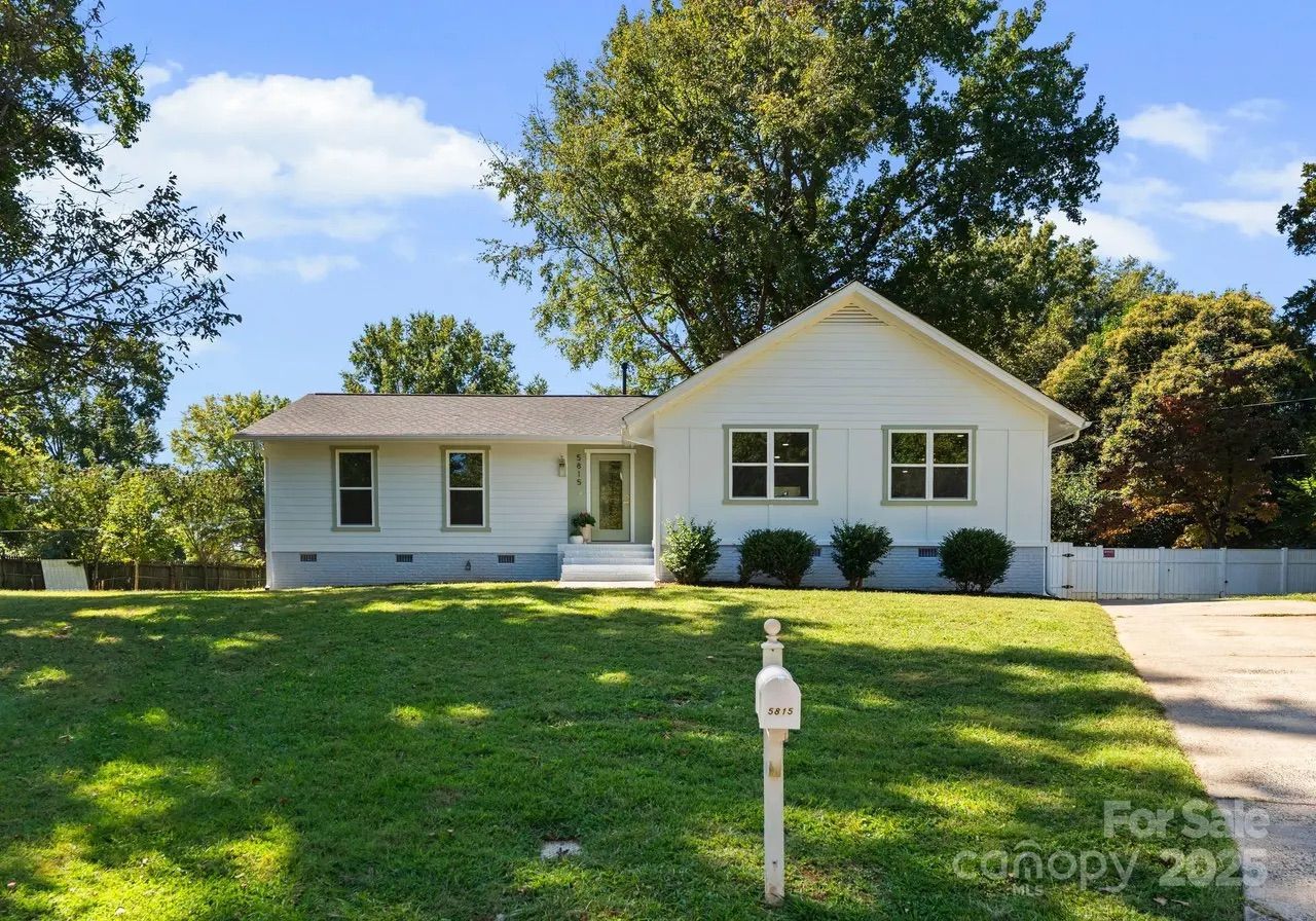 White single-story house with gray trim, green shrubs, and a mailbox labeled 5815 on a sunny day with a blue sky and trees in the background.