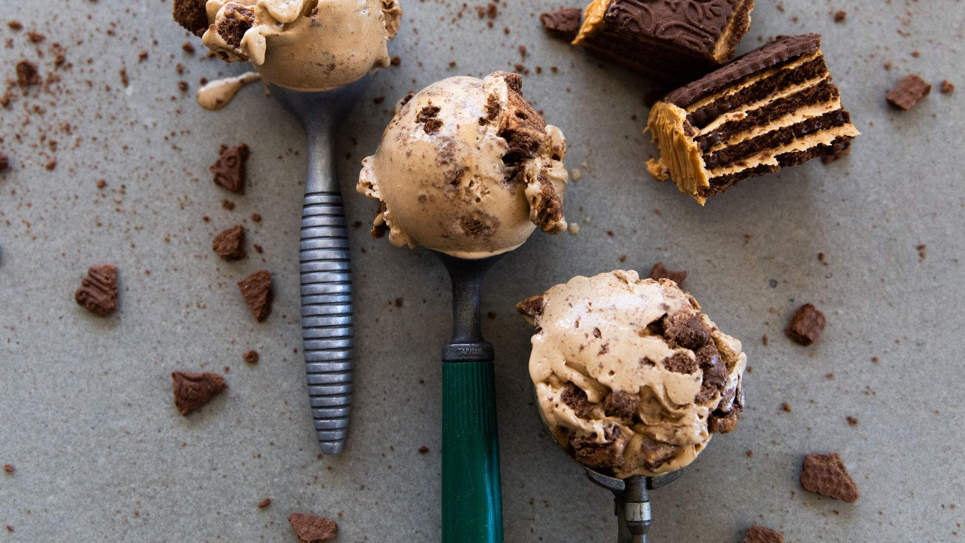 Top-down view of ice cream scoops with cookie pieces on spoons, chocolate wafer bars, and scattered crumbs on a gray surface.